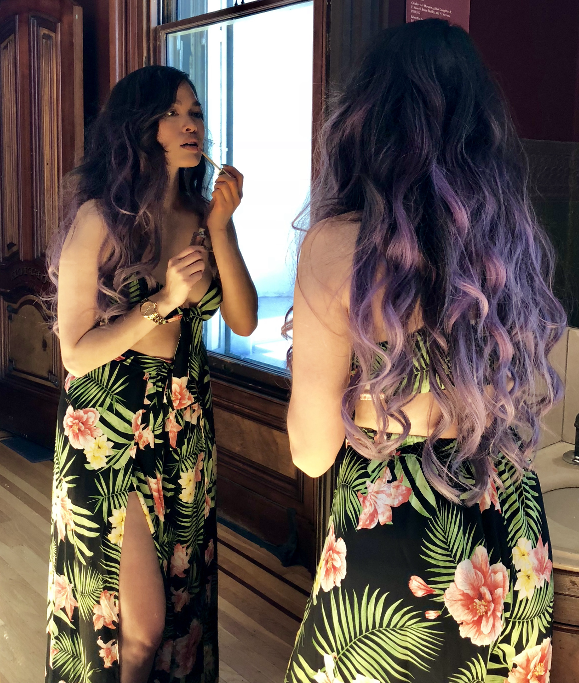 A woman with long, wavy purple hair stands in front of a mirror, applying lipstick, wearing a floral leaf-patterned dress with a slit.