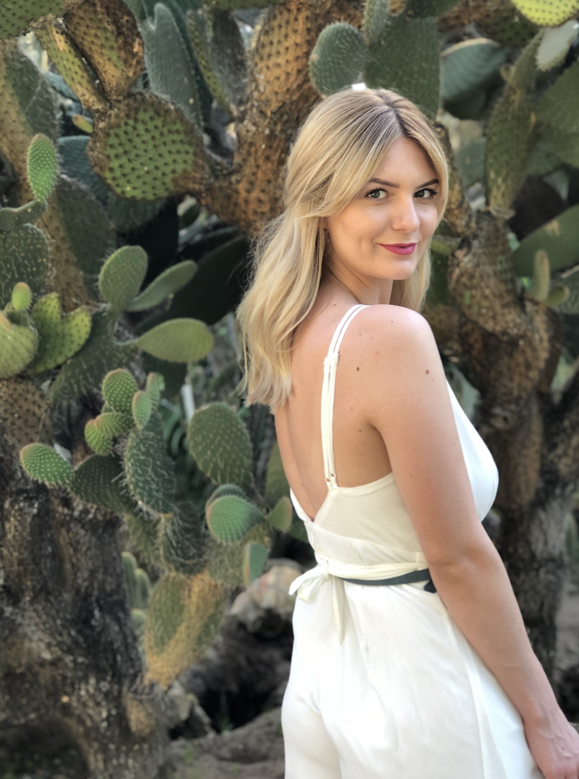 A woman with natural highlight blonde hair wearing a white dress standing in front of a large cactus plant.