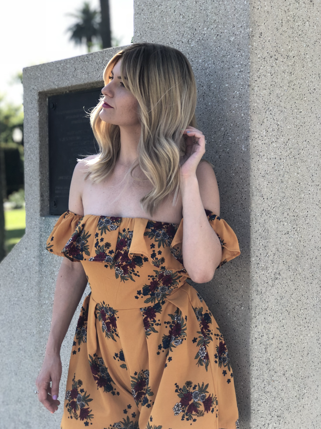 A woman with long blonde, wavy hair wearing an off-the-shoulder rust-colored floral dress standing outdoors against a concrete wall.
