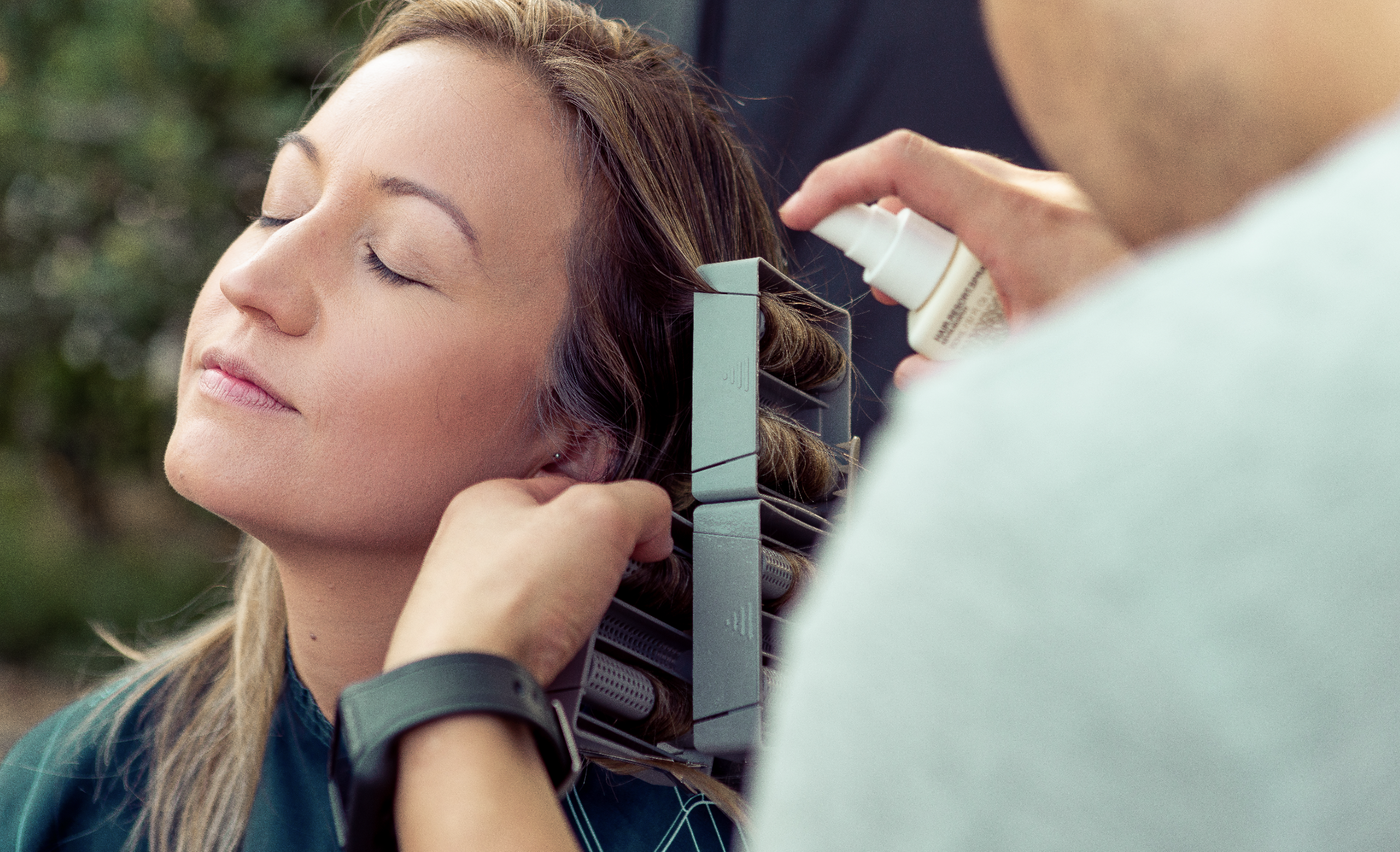 A woman is receiving a hair treatment or styling at a salon while her eyes are closed. A stylist is holding a spray bottle near her hair with what's likely a hair product. The woman has long blonde hair, and the background shows green foliage.