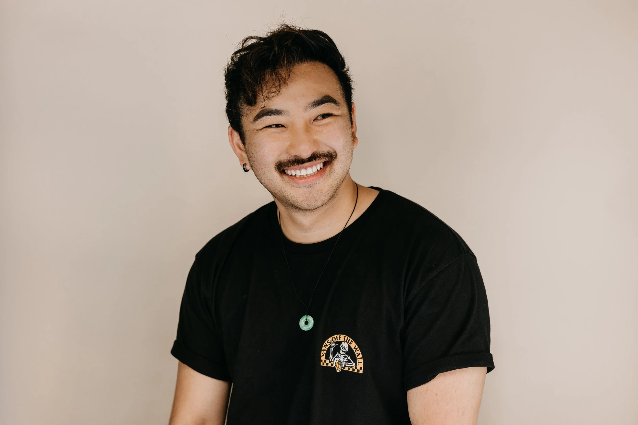 A young man with dark, wavy hair and facial hair, smiling while looking to the side, wearing a black t-shirt with Vans logo and a round green pendant necklace, against a plain beige background.