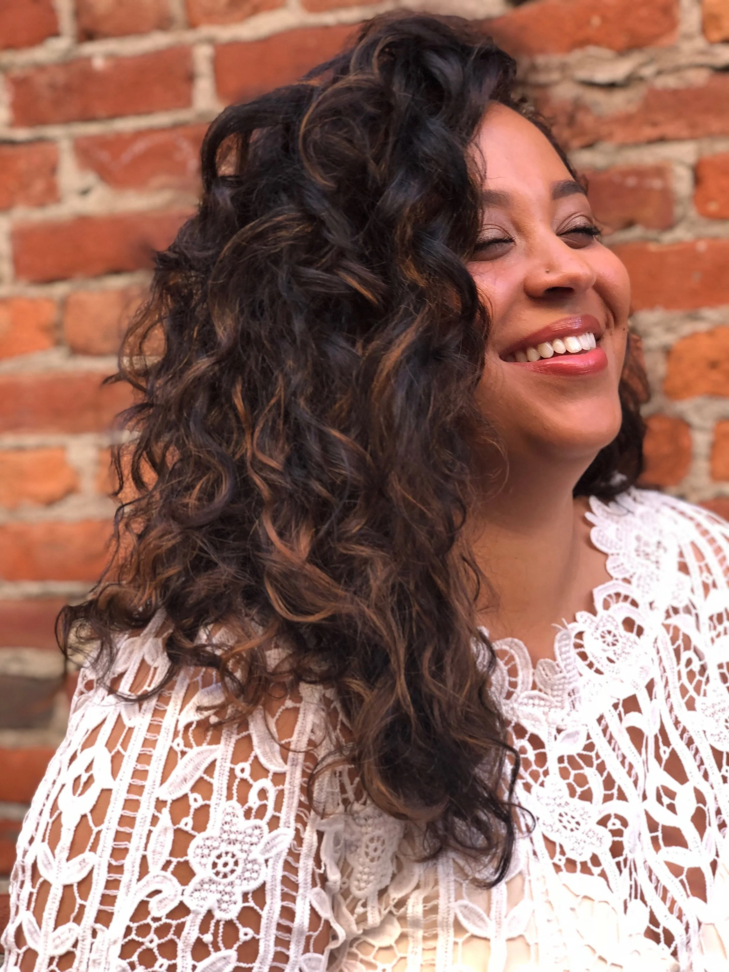 A woman with long, curly dark hair and light brown highlights, smiling, wearing a white lace top, standing against a brick wall.
