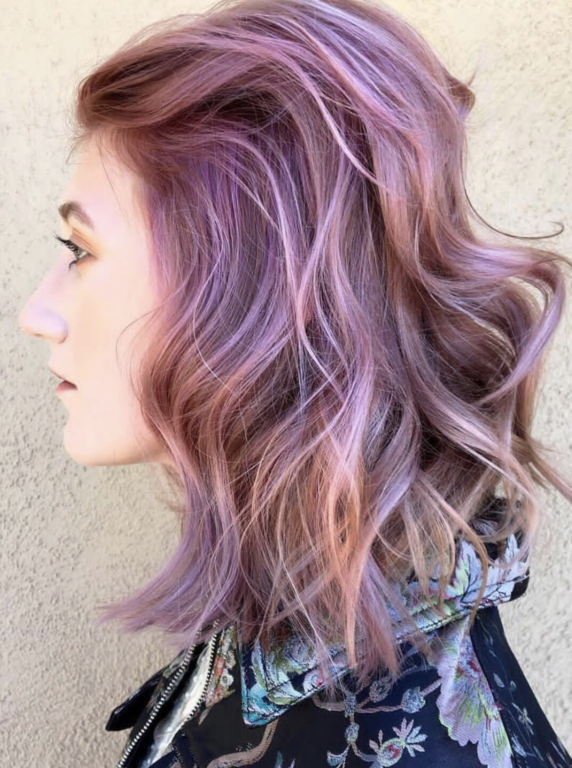 Side view of a woman with wavy, edgy pastel pink and purple hair, wearing a black floral embroidered jacket.