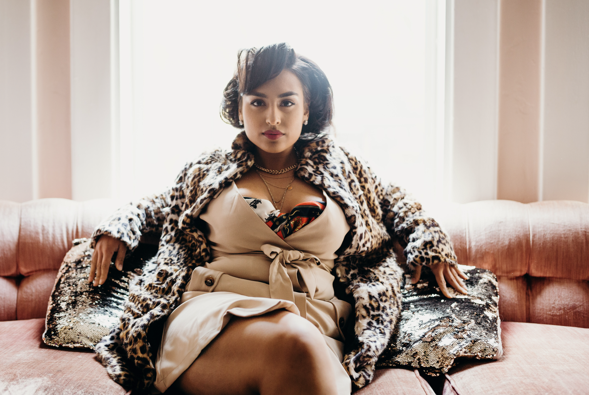 Woman sitting on a pink sofa with a leopard print coat, wearing a beige dress with a bow, accessorized with layered gold necklaces, and posing confidently.