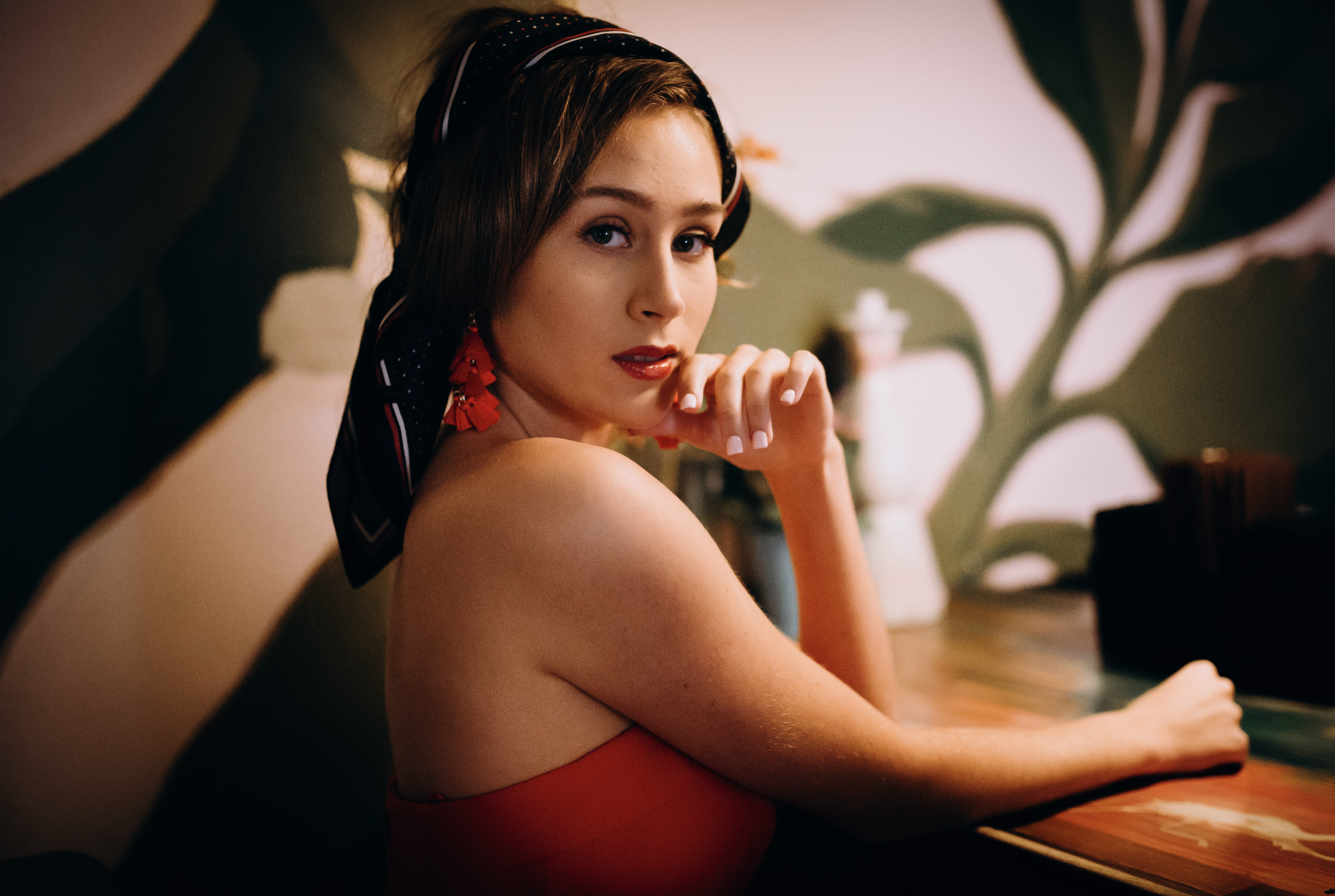 A woman with short hair, red lipstick, and red earrings, wearing a red strapless top and a black and red headscarf, sitting at a wooden table in a dimly lit room with floral wall art.