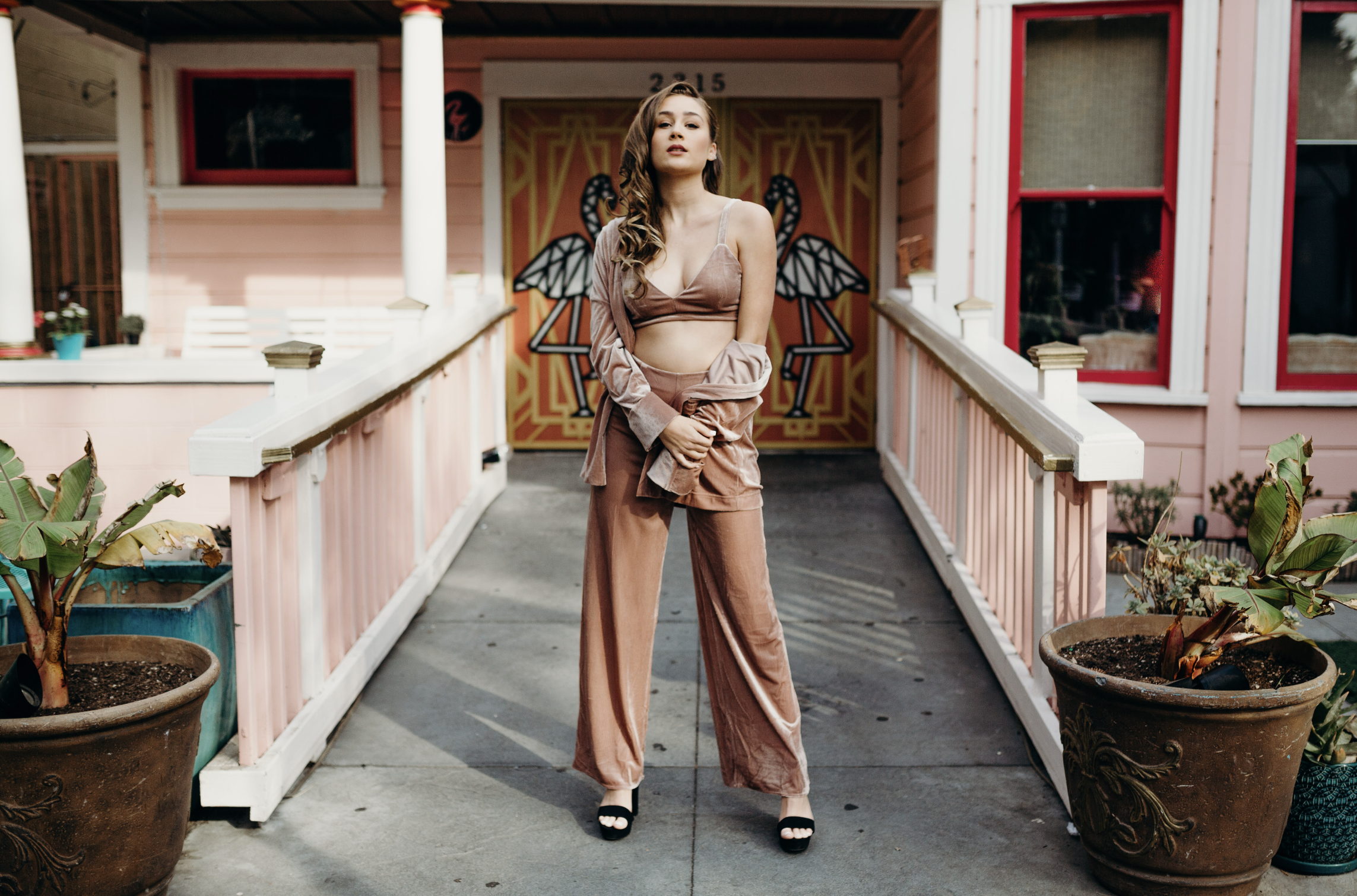 A young woman with long, wavy hair standing on a porch in front of a colorful door with a swan painting. She wears a brown crop top, matching wide-leg pants, high heels, and a light jacket draped over her shoulders.