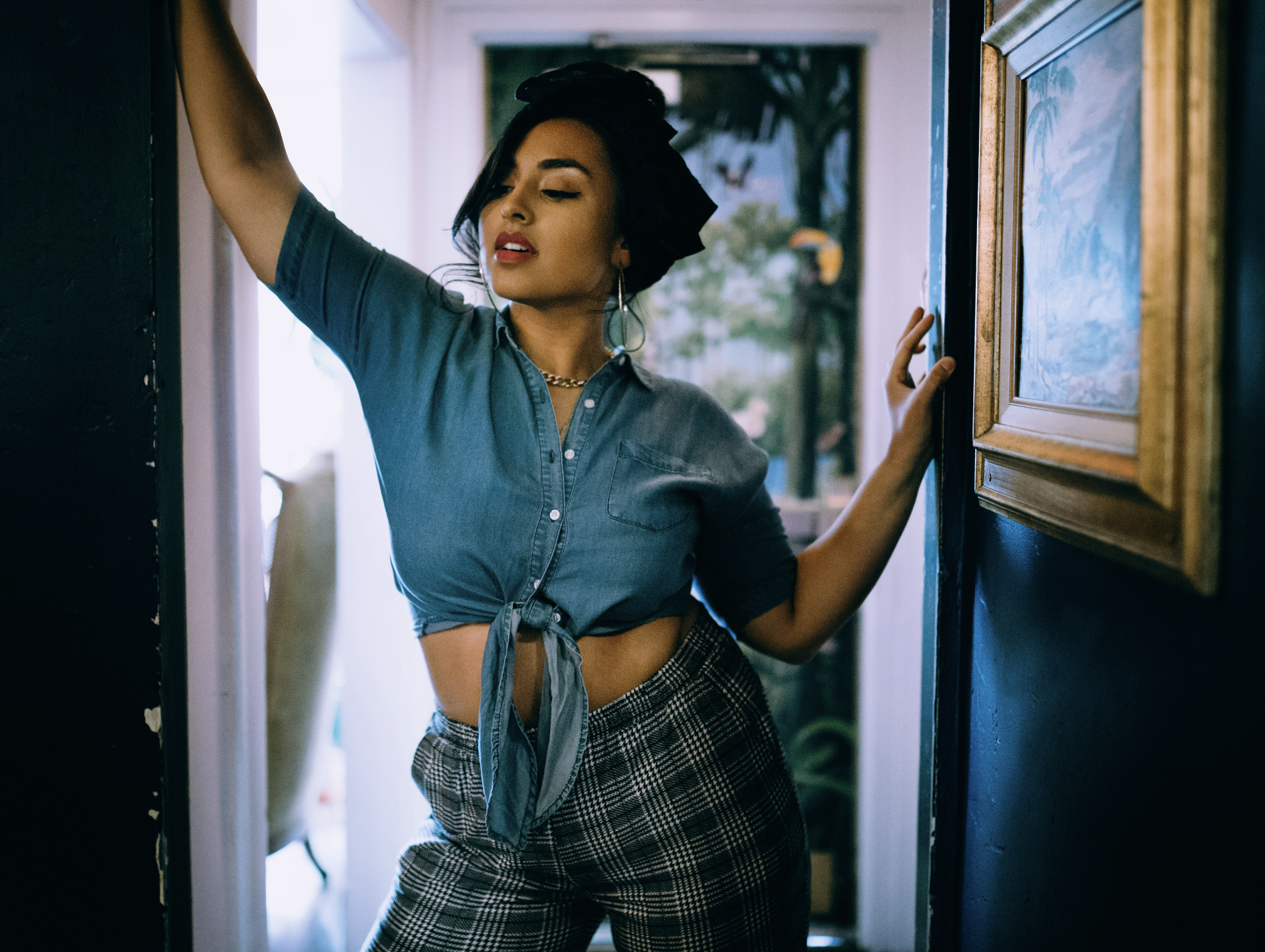 A photoshoot of a woman in a tied denim shirt and plaid pants poses with one arm raised and her eyes closed in a well-lit room with artworks and a window showing outdoor scenery.