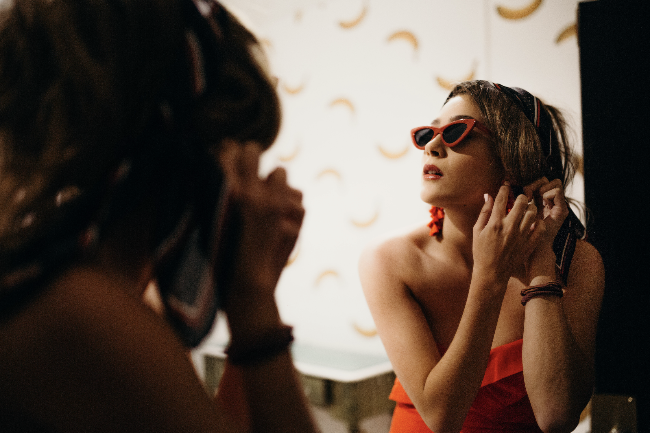 A woman wearing red sunglasses, red lipstick, and large earrings adjusting her ear in front of a mirror, in a room with a patterned wallpaper.