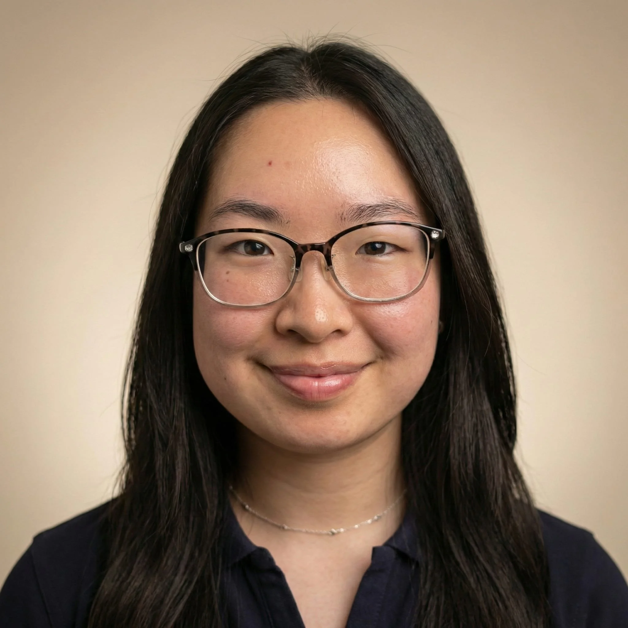 Portrait of a smiling woman with glasses, long black hair, and wearing a black top and a delicate necklace, against a plain beige background.