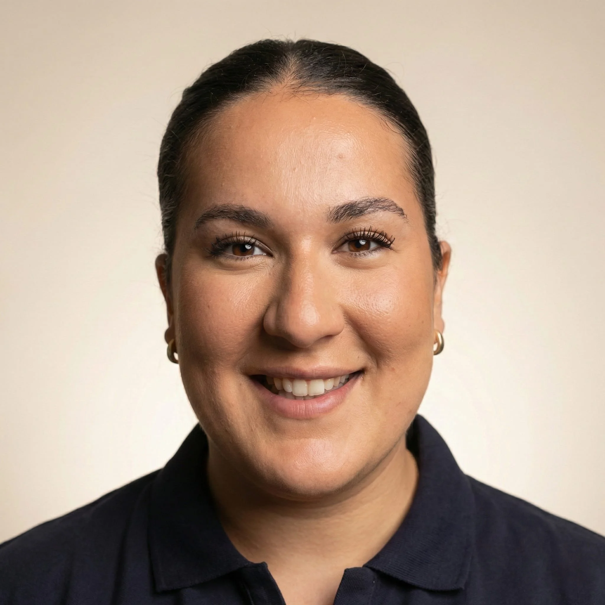 A smiling woman with dark hair slicked back, wearing small hoop earrings and a dark collared shirt, standing against a neutral background.