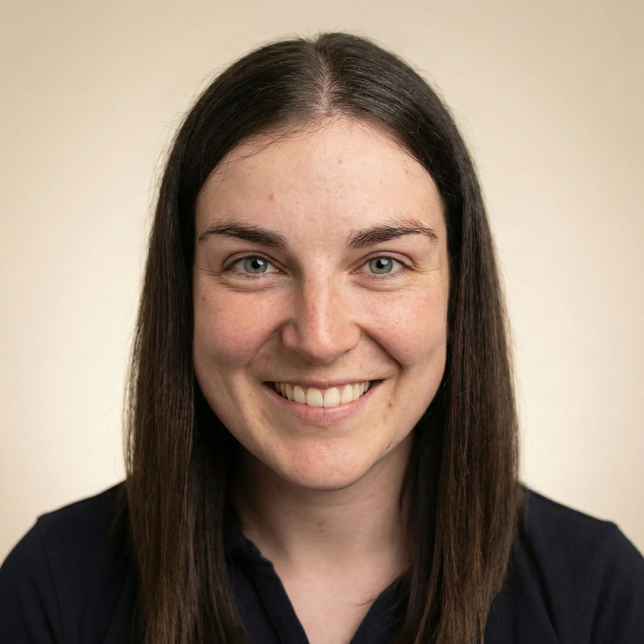 Close-up portrait of a woman with long brown hair, blue eyes, smiling, wearing a black shirt, against a plain beige background.