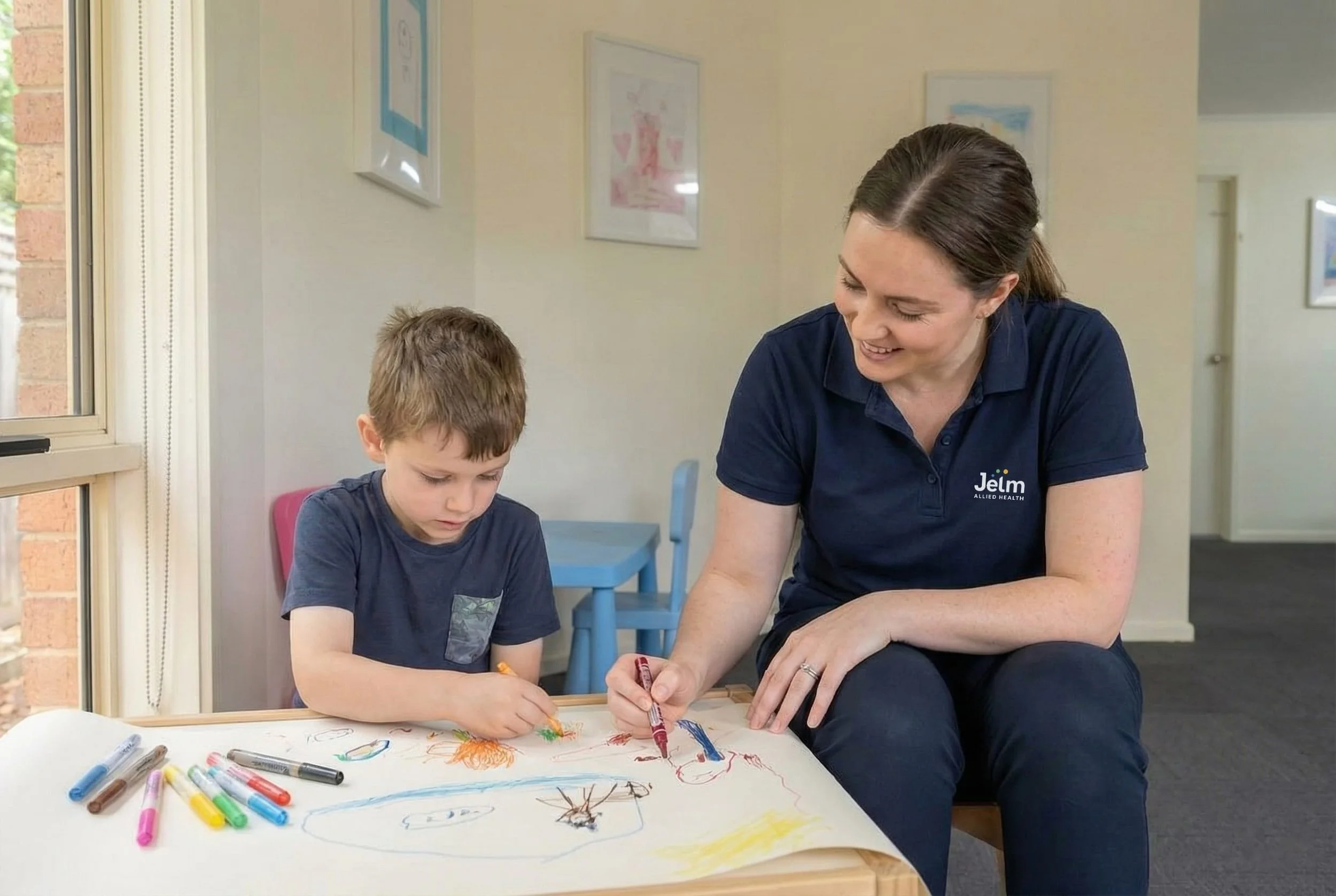 A woman and a young boy are sitting at a table drawing with colored markers. The woman is smiling, wearing a navy polo with the logo 'Jelm Allied Health'. The boy is focused on his drawing, which includes various colorful sketches, and there are multiple markers on the table.