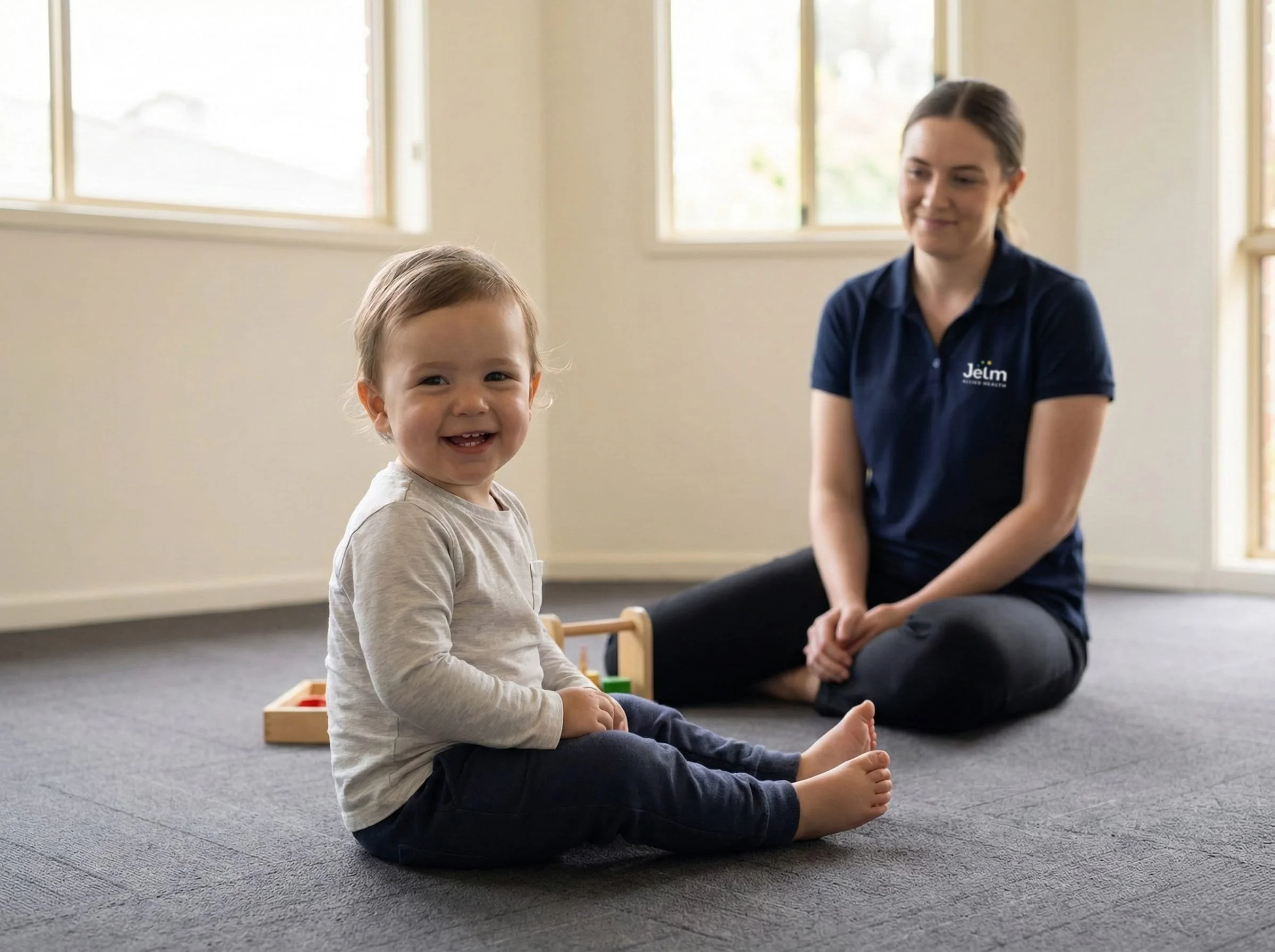 A young child with a big smile, sitting on a carpeted floor with legs crossed, in a room with large windows, and a woman sitting nearby, smiling and watching the child.