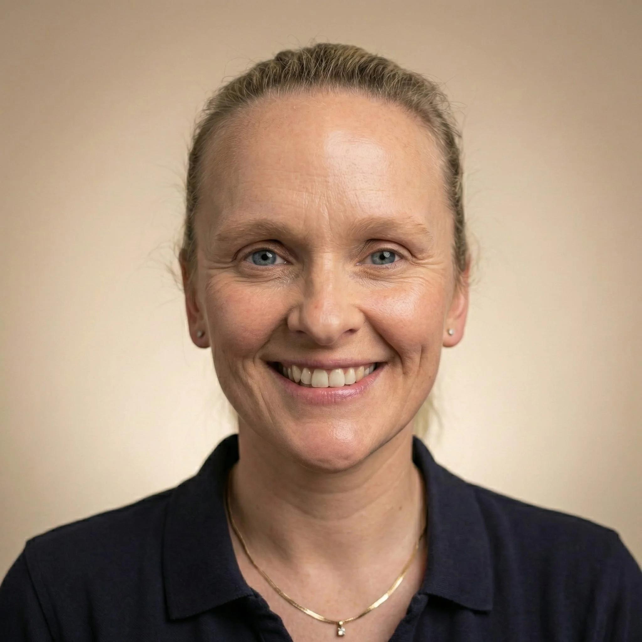 Close-up portrait of a smiling woman with blonde hair tied back, wearing a dark collared shirt and a delicate gold necklace.