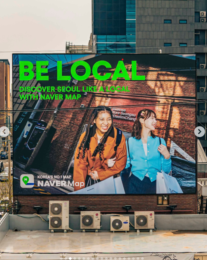 Large outdoor digital billboard advertising Naver Map with the text 'BE LOCAL' and 'Discover Seoul like a local with Naver Map' showing two smiling women walking in Seoul, South Korea, carrying shopping bags, with buildings and air conditioning units below.