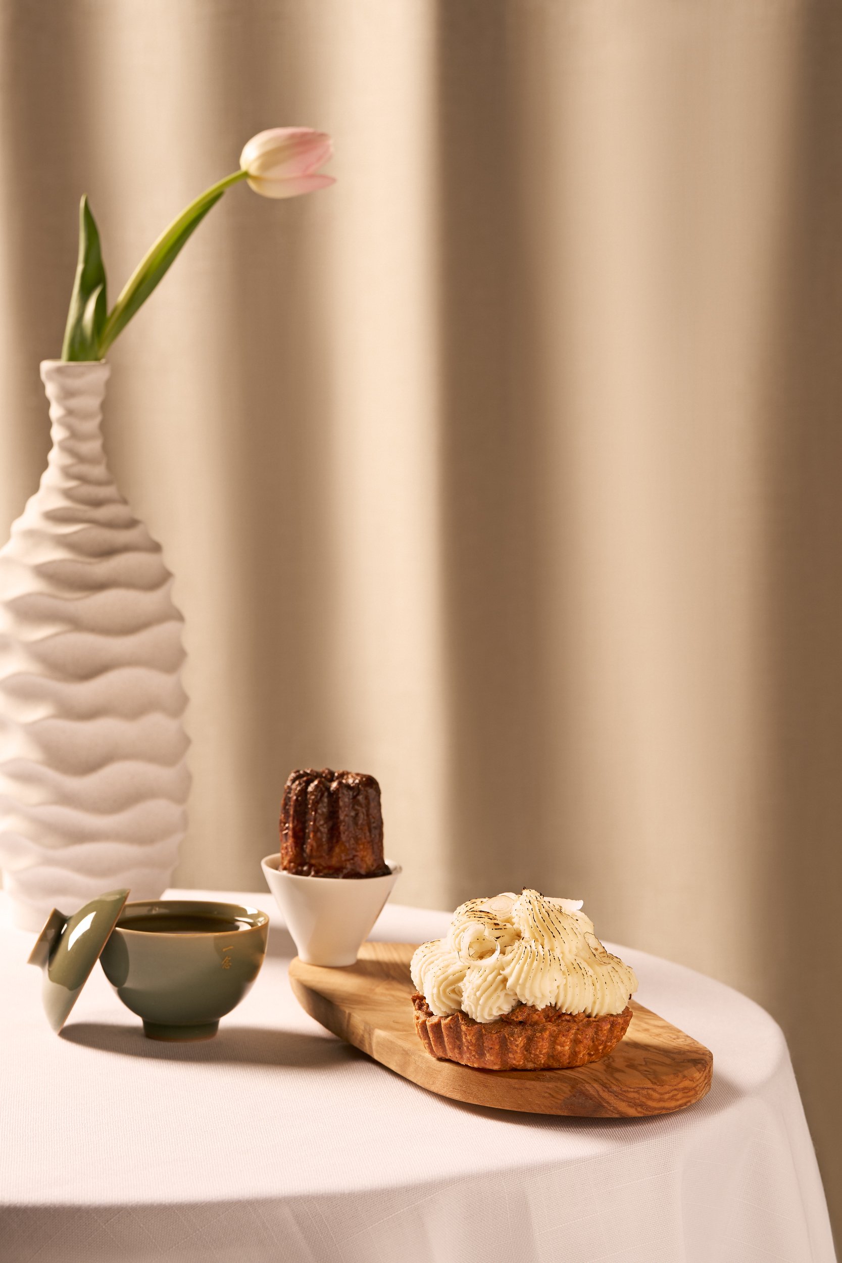 A table with a green teacup filled with a dark beverage, a small loaf cake with chocolate glaze in a white cup, and a tart with white cream frosting on a wooden board, next to a tall white vase with a pink tulip.