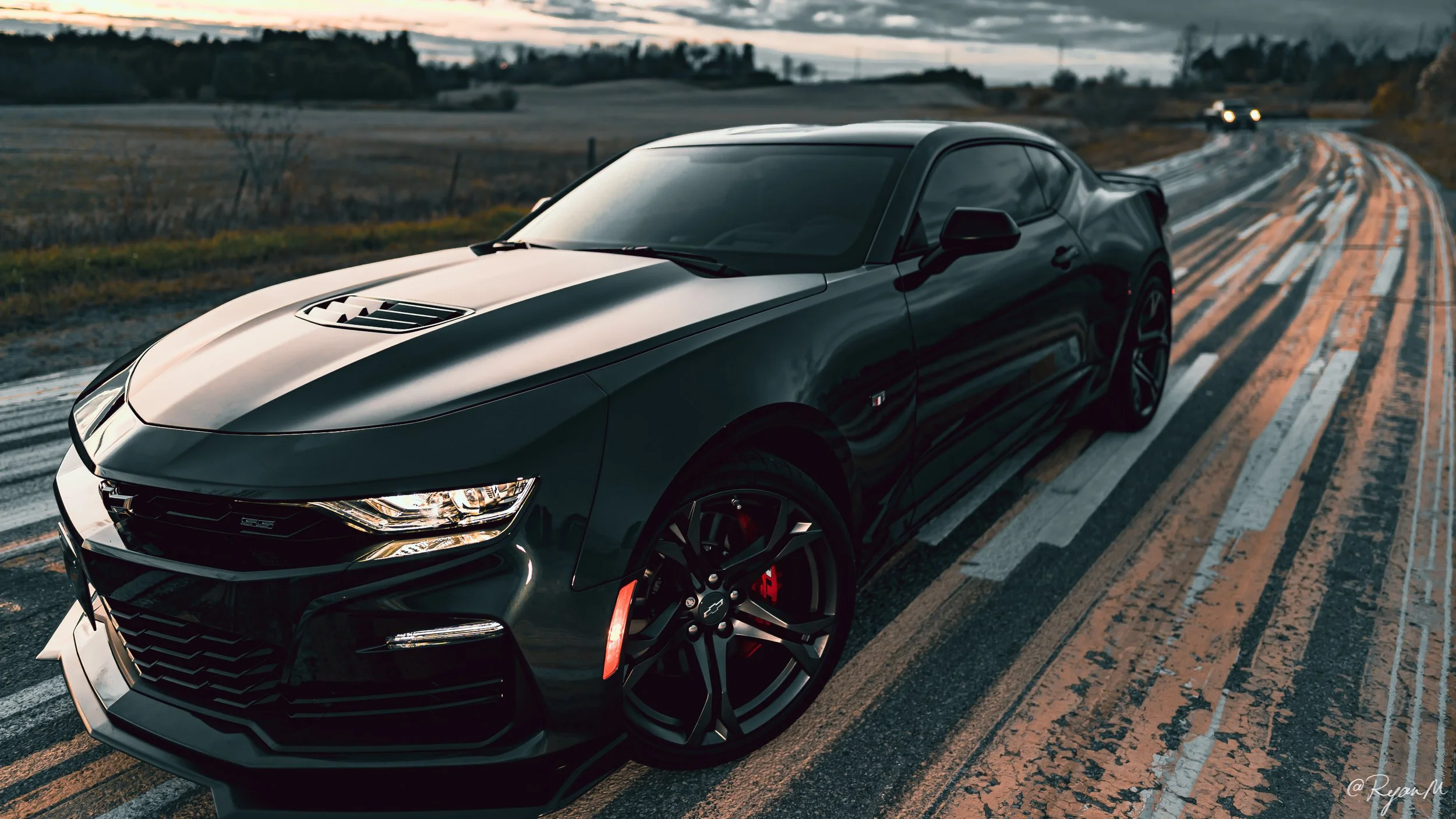 A sleek black sports car with a hood vent, sharp headlights, and black rims with red brake calipers, parked on a wet road at sunset, reflecting the fading light.