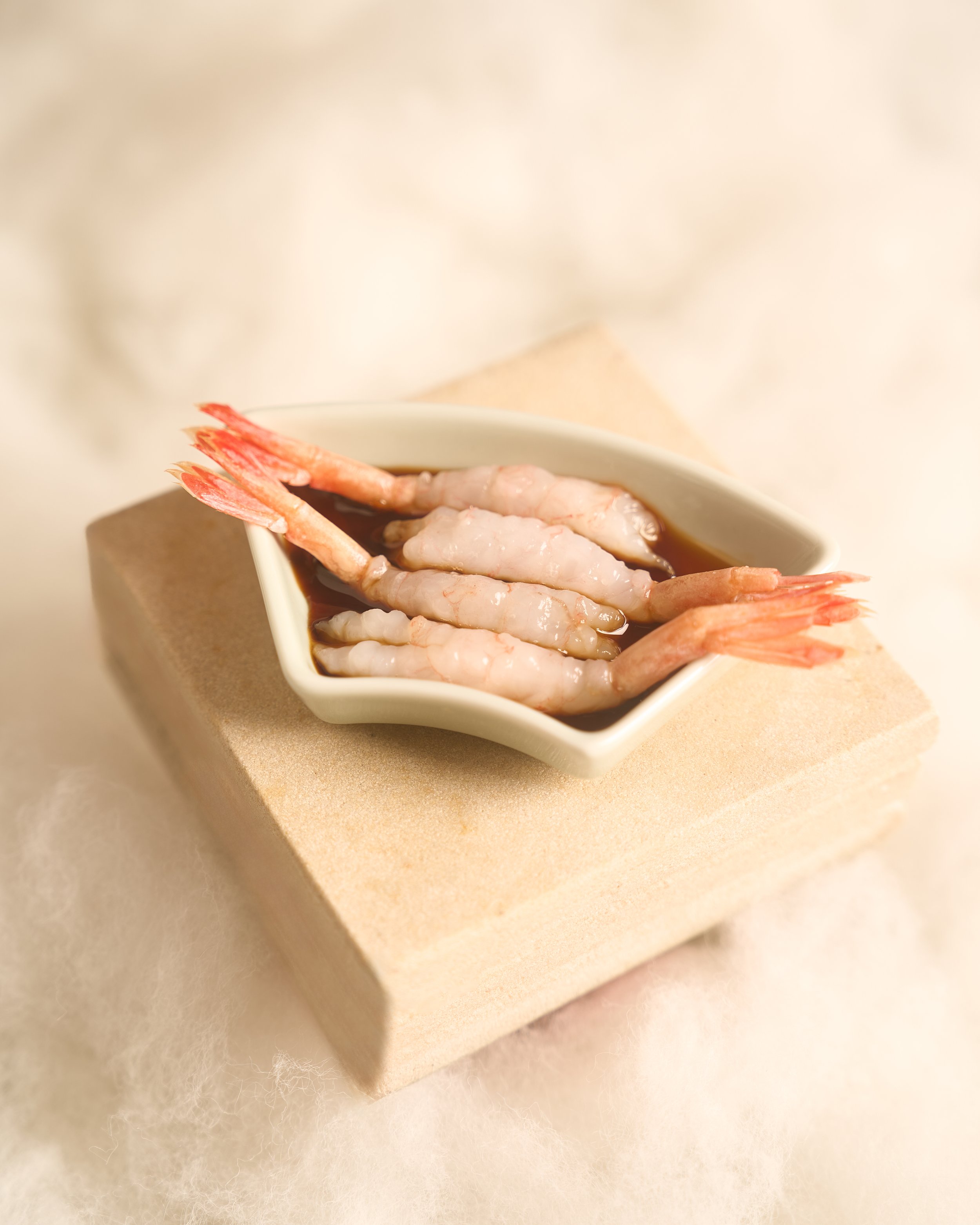 Close-up of a small square dish filled with cooked shrimp in a dark sauce, placed on a beige surface.
