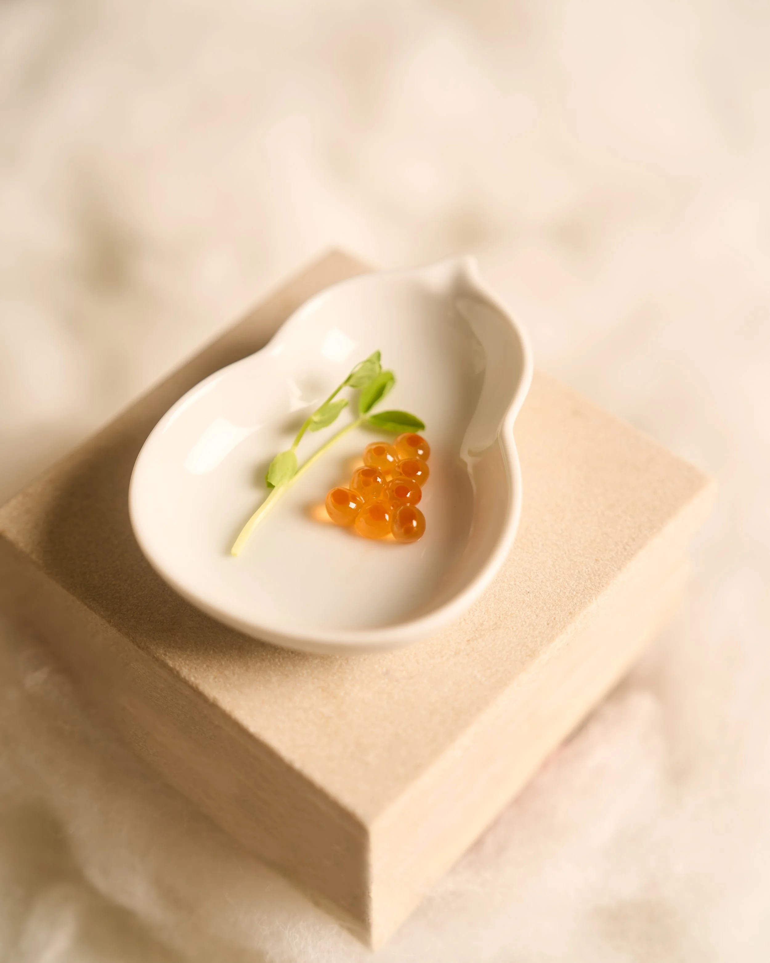 Close-up of a white ceramic dish with orange salmon roe, small green leaves, and a tiny piece of fish on a beige surface.