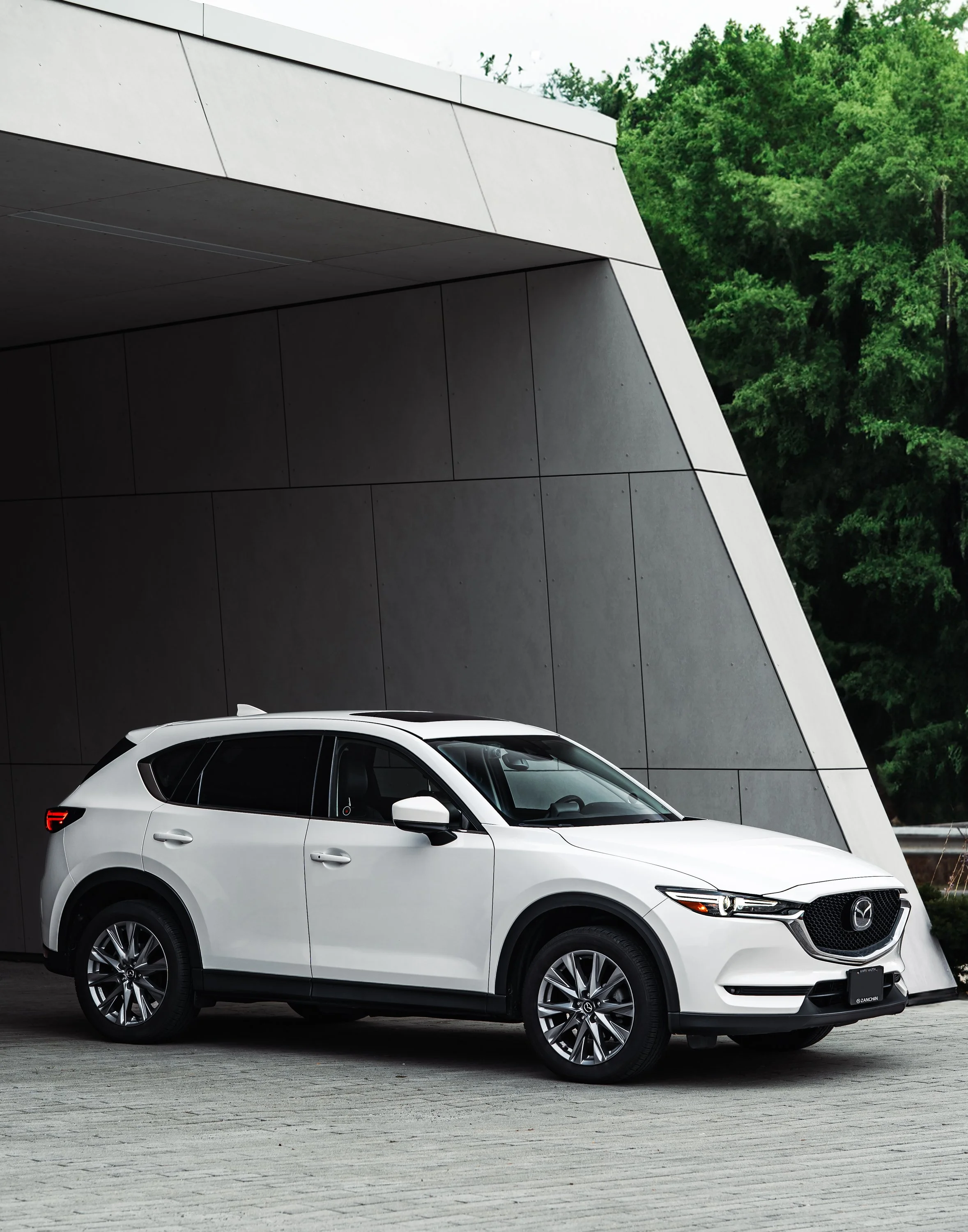White Mazda SUV parked in front of a modern building with a large overhang, green trees in the background, and a paved area in the foreground.
