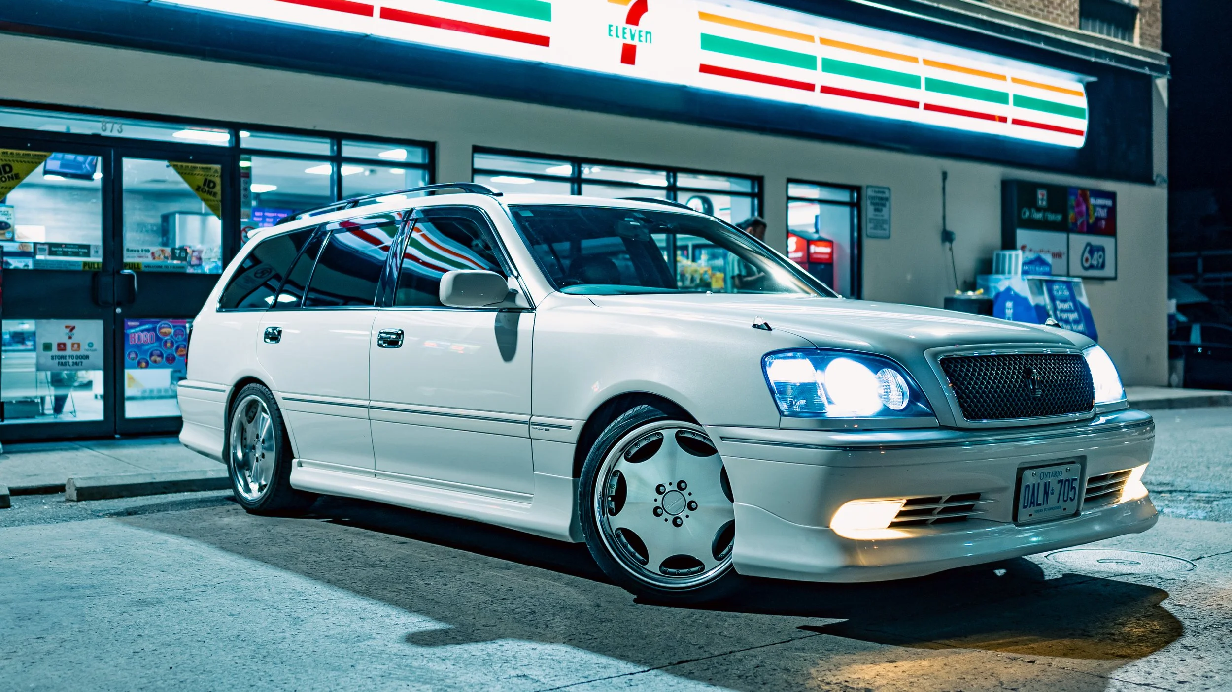 White luxury sedan parked outside a 7-Eleven convenience store at night, with store signage and windows visible in the background.