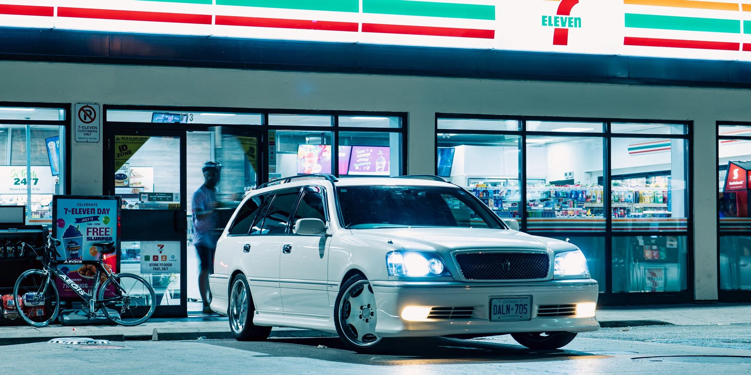 A white luxury sedan car is parked in front of a 7-Eleven convenience store during nighttime, with its headlights on. The store has a large illuminated sign and glass windows showing inside, with bicycles parked outside and promotional signs near the