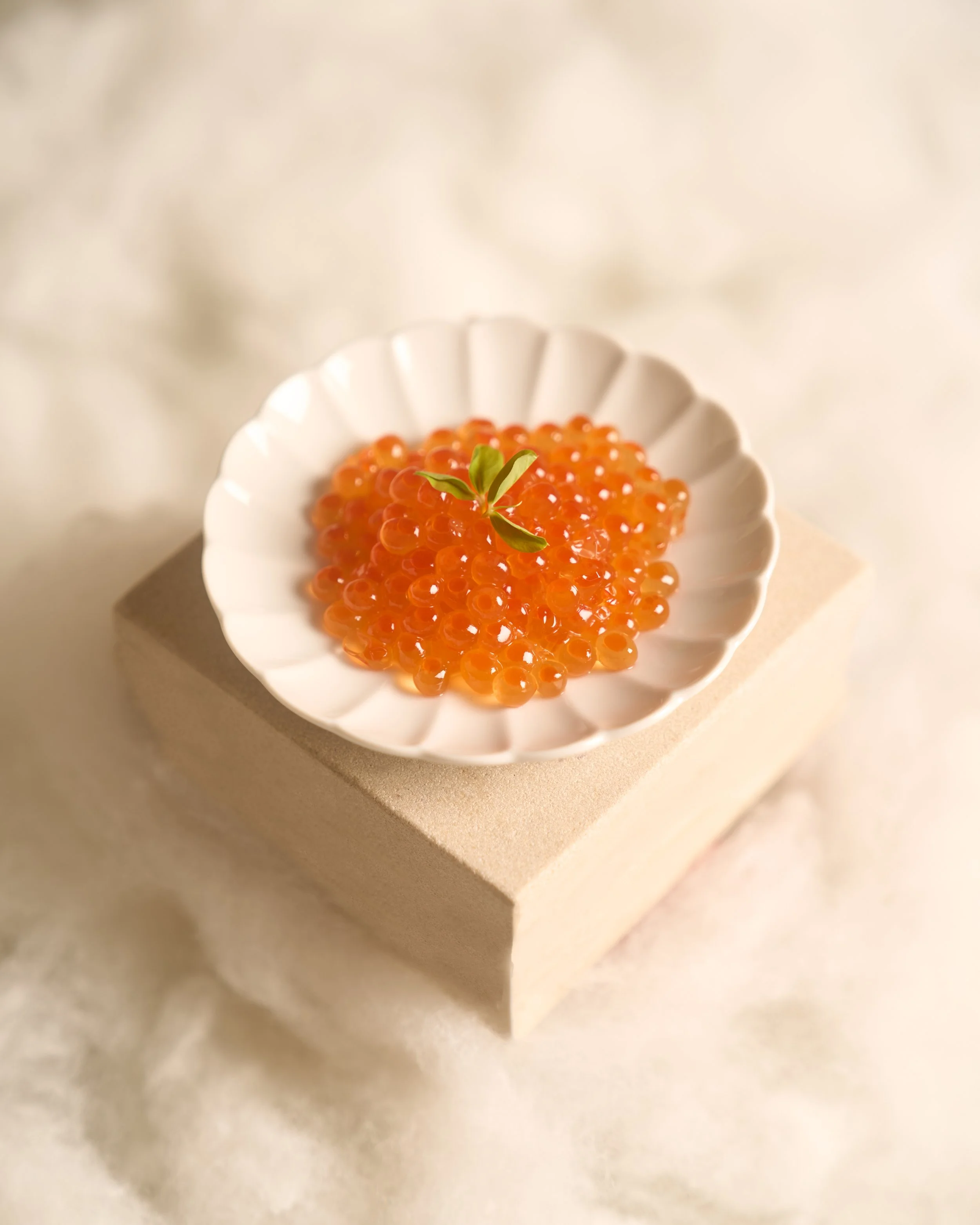 A small white dish filled with bright orange salmon roe, garnished with a small green leaf, on a beige surface with a blurred background.