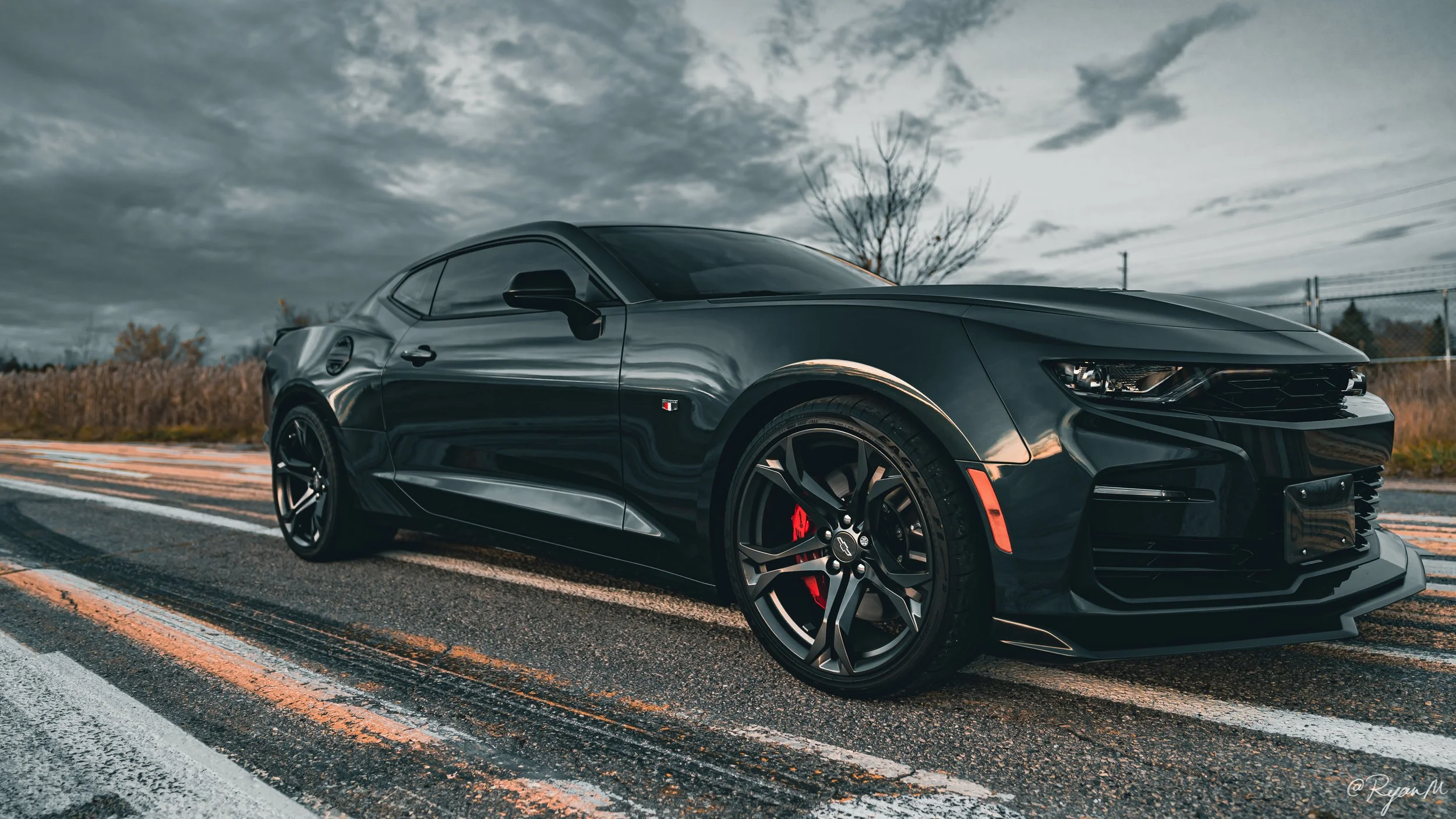 Black sports car parked on the side of an empty road under a cloudy sky, with autumn trees and a chain-link fence in the background.