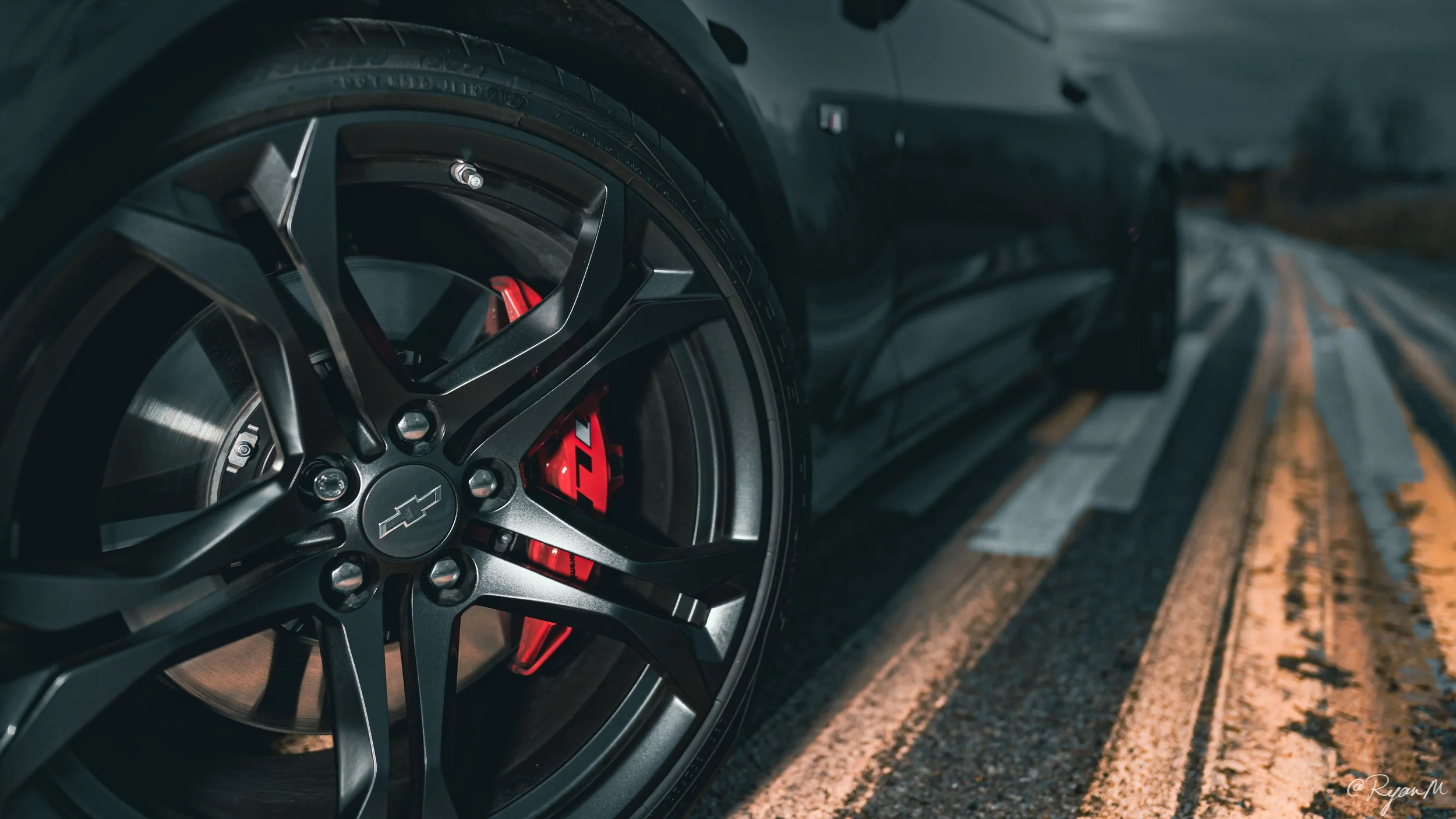 Close-up of a black car wheel with a Chevrolet logo on the center cap, red brake caliper behind the wheel, on a snowy road with yellow and blue lines.