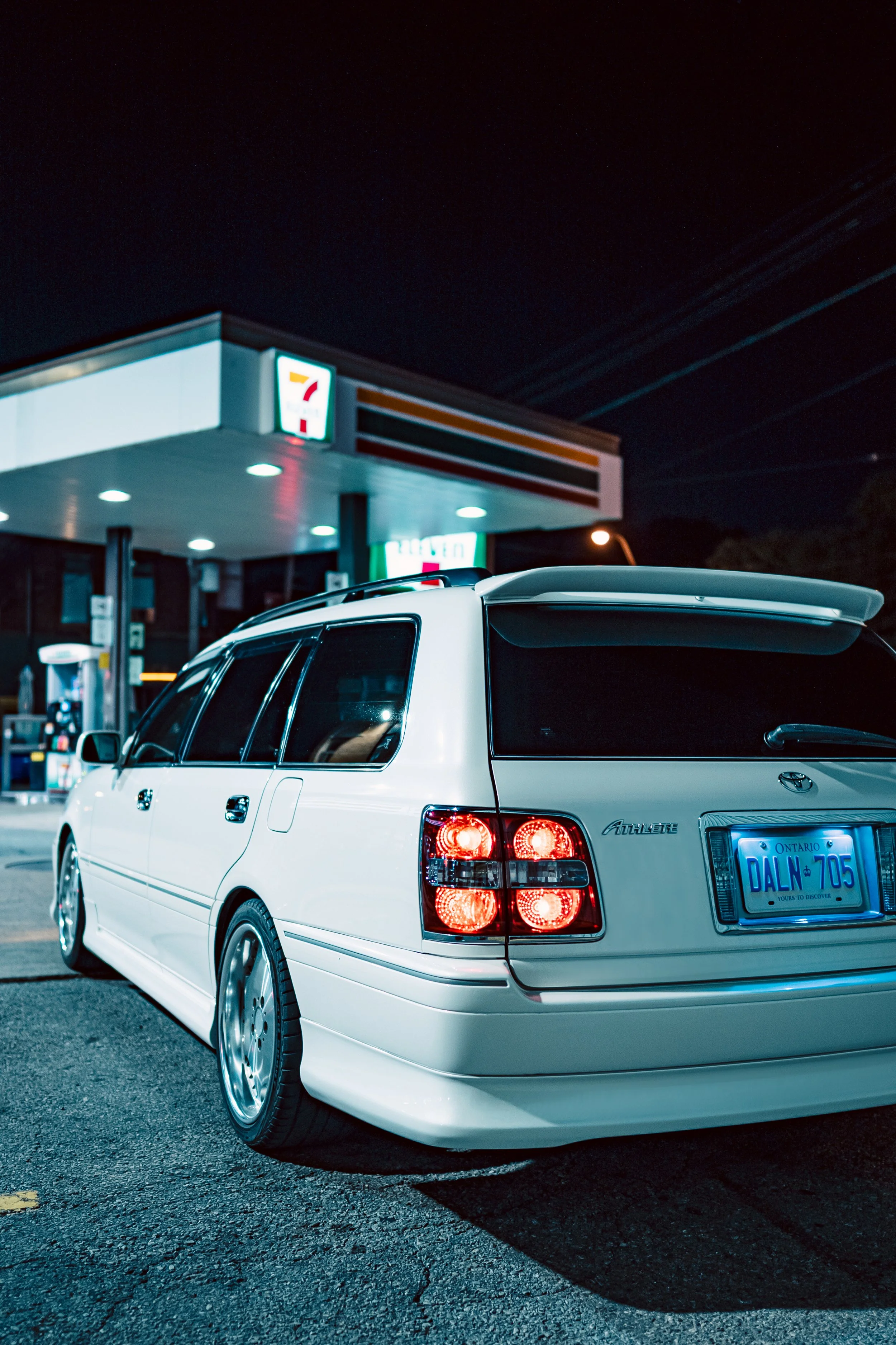 A white Toyota Prius parked in front of a 7-Eleven convenience store at night, with the store's illuminated sign visible in the background.