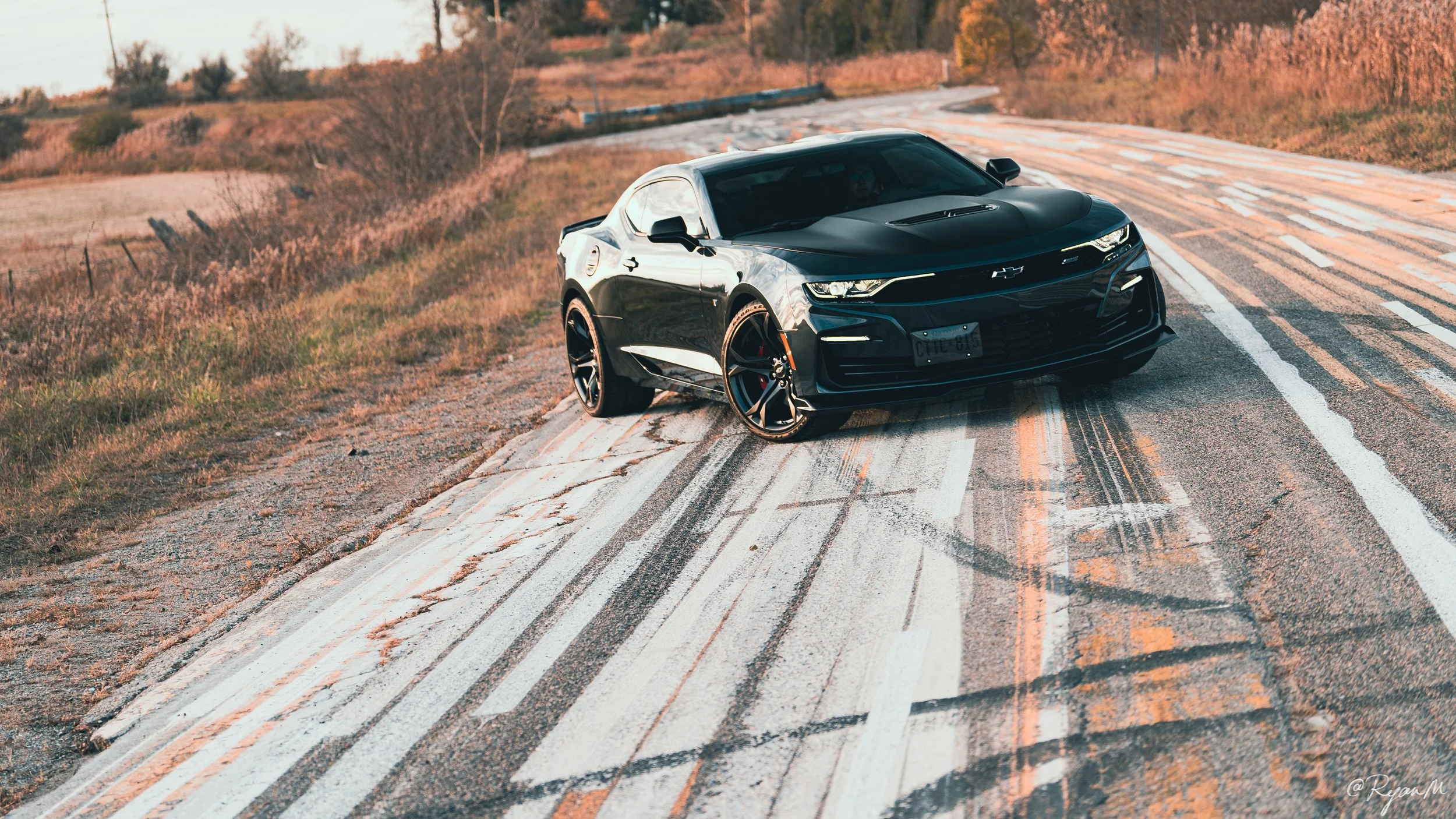 A black sports car with a sleek design parked on a damaged rural road with cracks and faded white lines, surrounded by dry grass and sparse trees in the background during sunset.