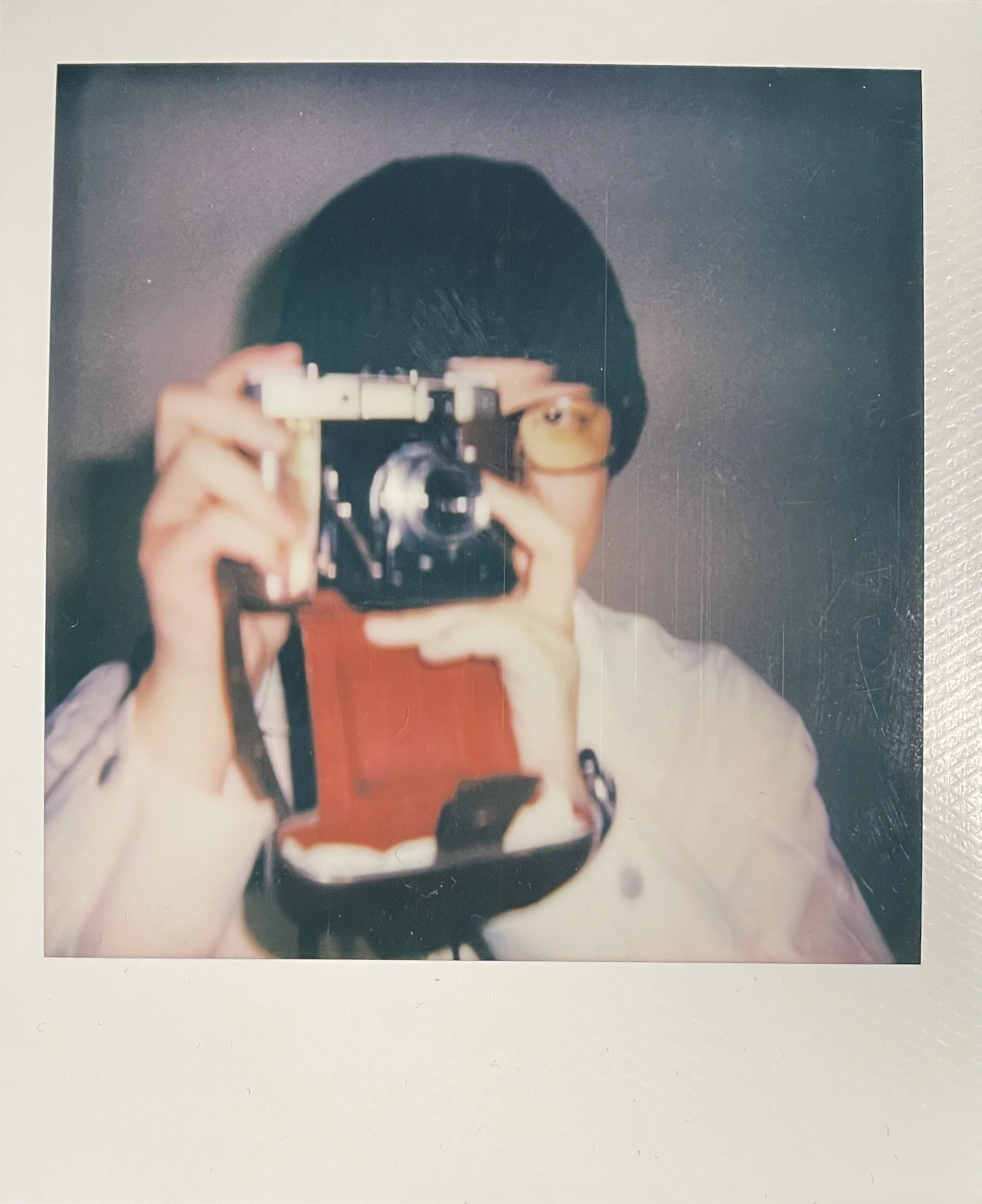 A person taking a mirror selfie with a vintage camera, wearing a tan hat and a white shirt with a red vest, against a plain dark background.