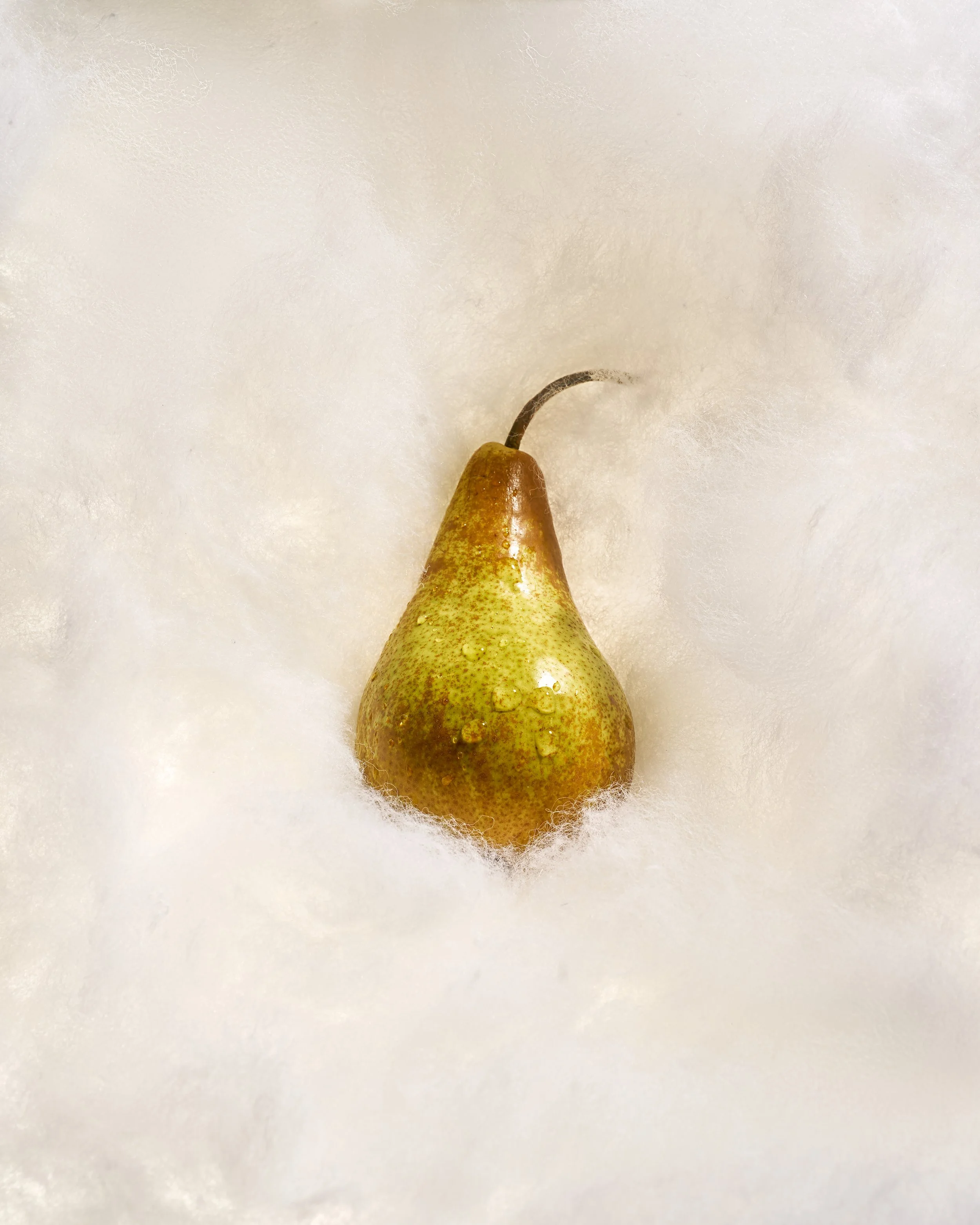 Close-up of a ripe pear with water droplets on its yellow-green skin, placed on a white textured surface.