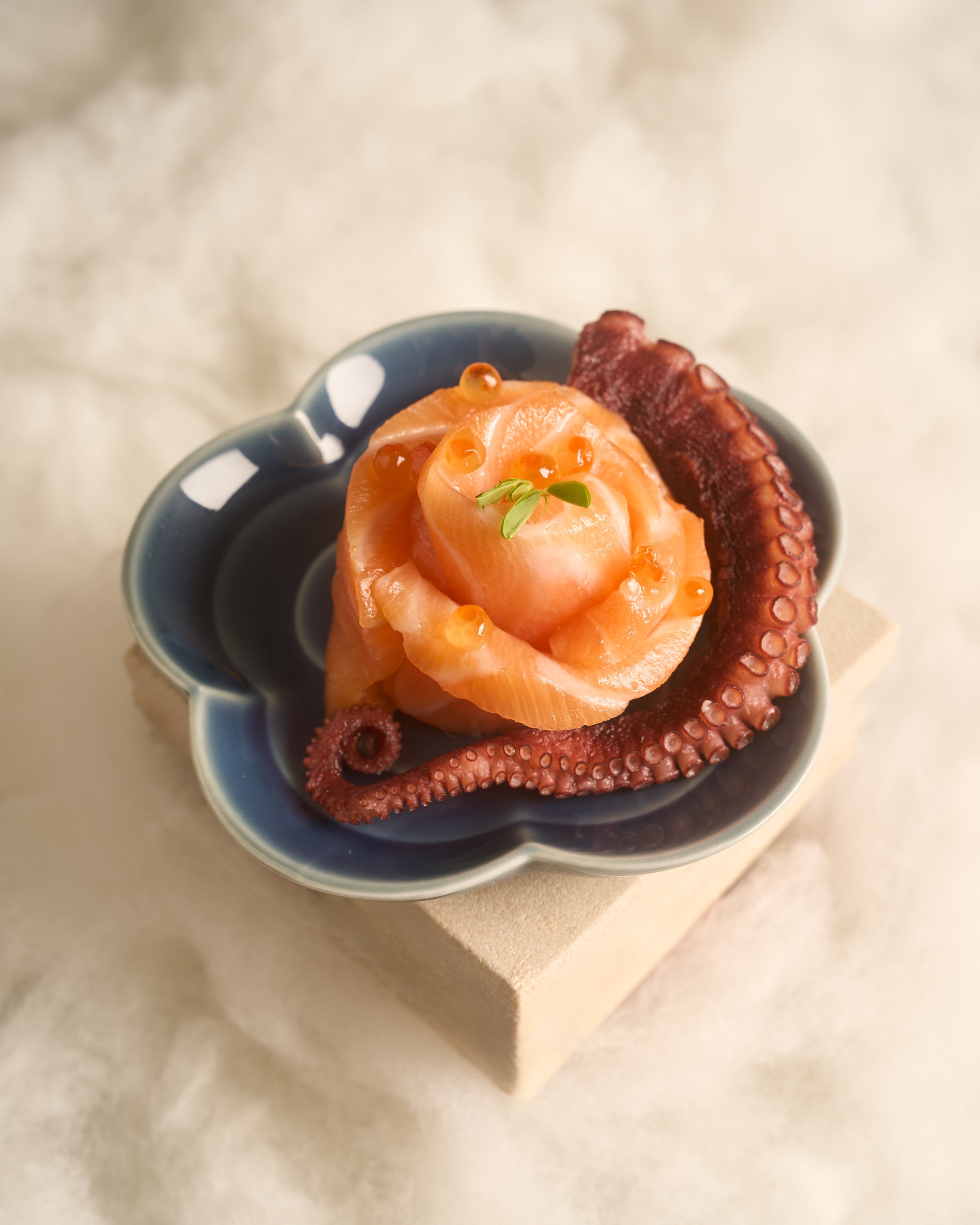 Close-up of a bowl containing smoked salmon, octopus tentacle, salmon roe, and a small green garnish. The bowl is placed on a light-colored surface.