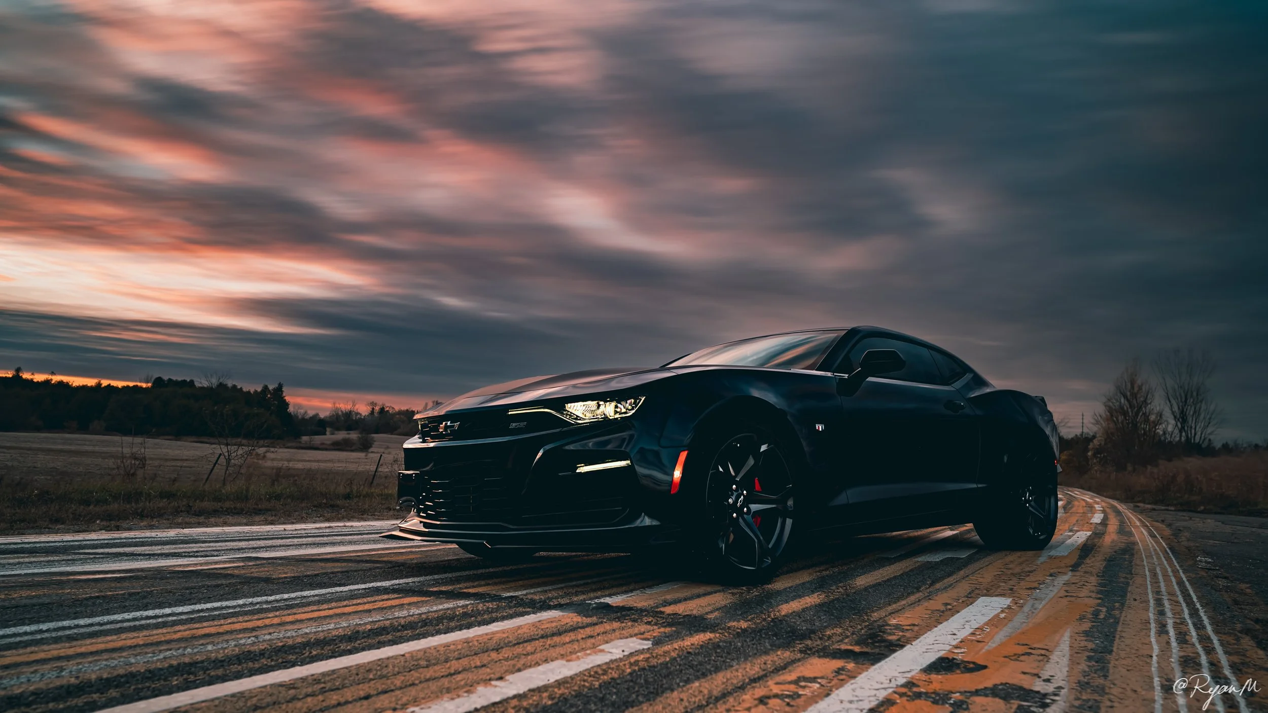 A black sports car parked on a rural highway at sunset with colorful clouds in the sky and trees in the background.
