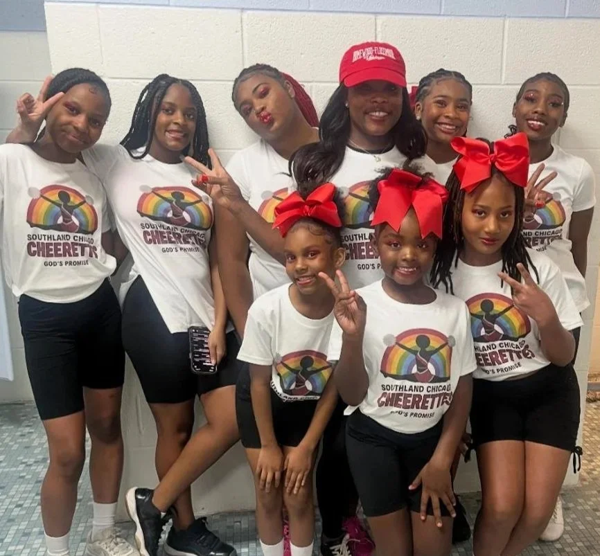 Group of eight girls wearing matching cheerleading T-shirts with a rainbow and cheerleader logo, some with large red bows in their hair, posing in a hallway with a white brick wall behind them, smiling and making peace signs.
