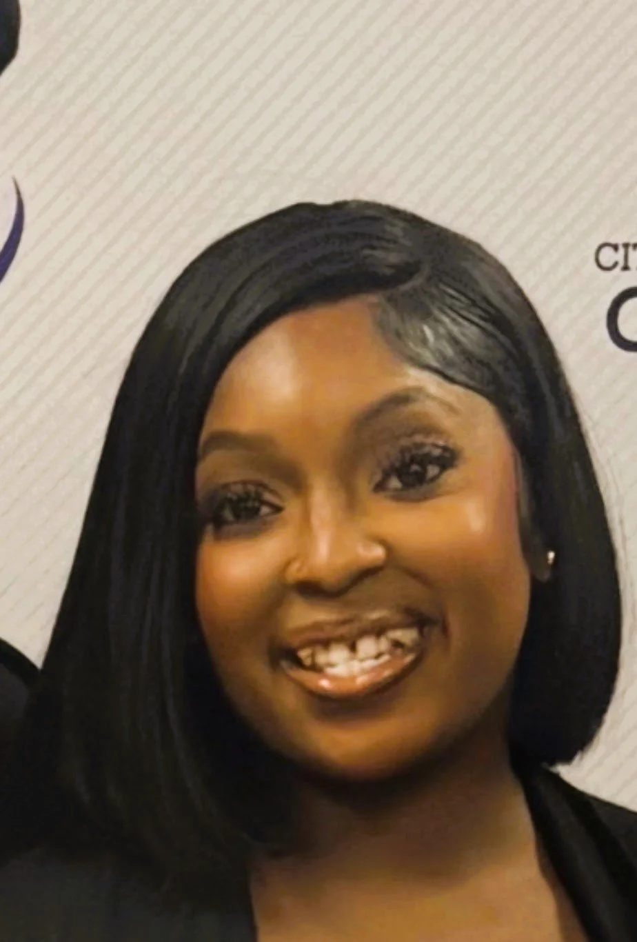 Close-up portrait of a woman with shoulder-length black hair, smiling, against a light textured background with part of a logo visible.