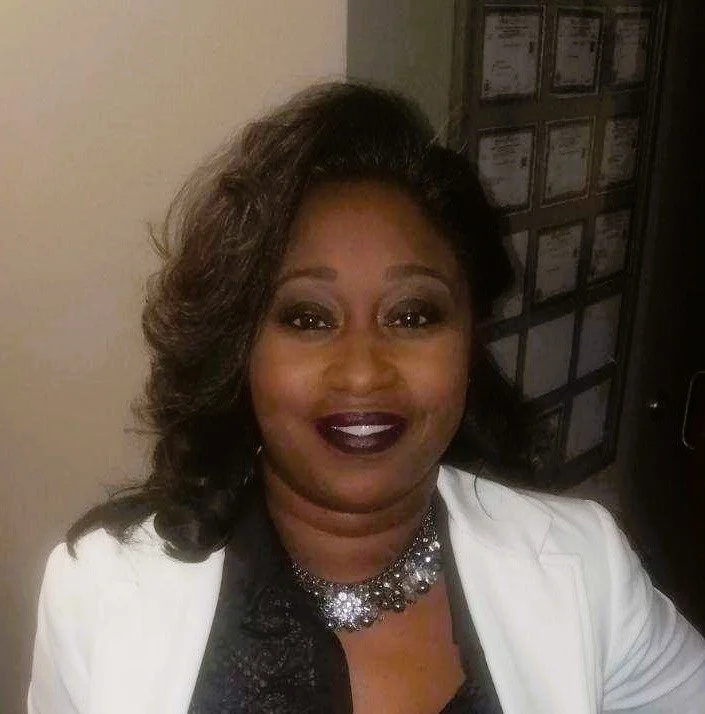 A woman with dark brown, shoulder-length hair styled in loose waves, wearing a white blazer, a black top, and a sparkling silver necklace, smiling at the camera.