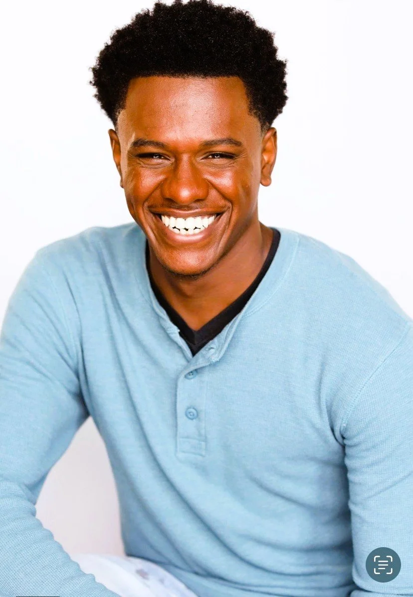 A smiling man with dark skin, short curly black hair, wearing a light blue shirt, sitting against a plain white background.