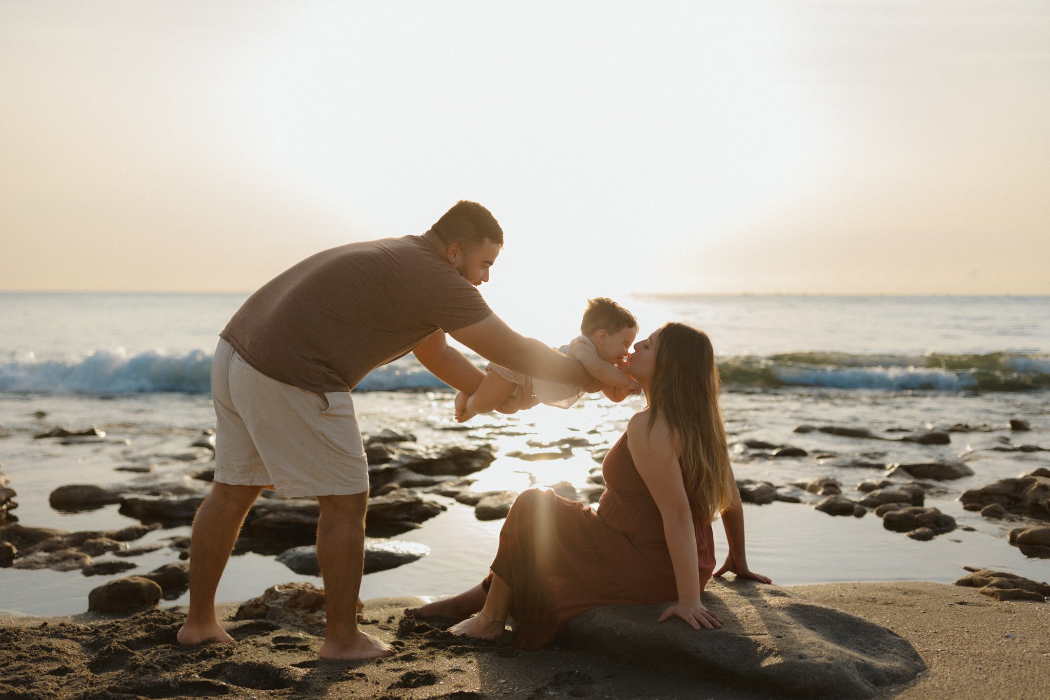 A family of three playing on the beach at sunset. The father is lifting the child towards the mother, who is sitting on a rock, as they smile at each other.