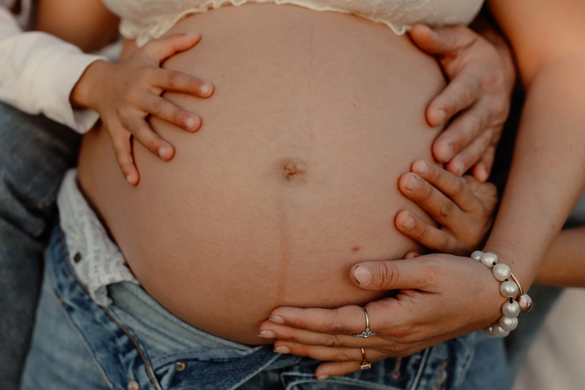 Close-up of a pregnant belly with multiple hands gently holding and supporting it, showing a mix of different skin tones and jewelry, including rings and a pearl bracelet.