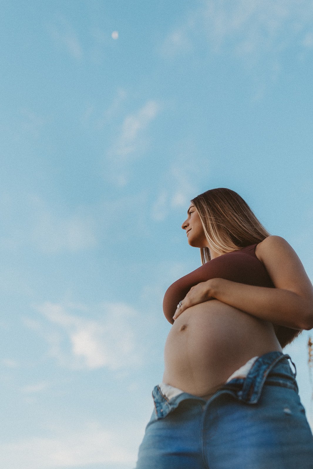 A pregnant woman standing outdoors during daytime, smiling and holding her belly, with a blue sky and clouds in the background.