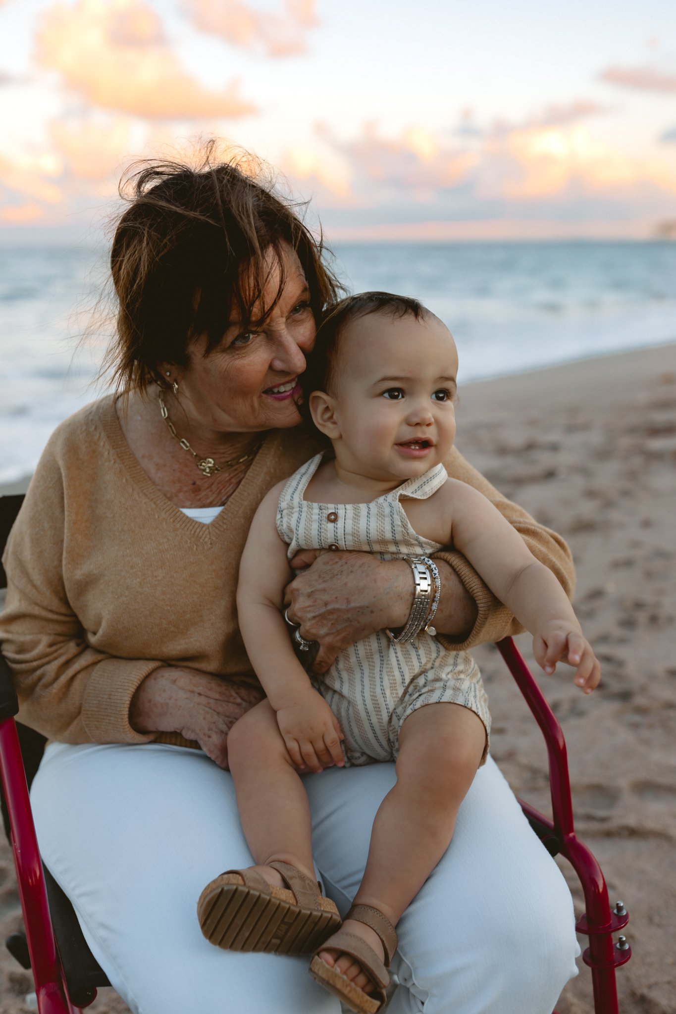 An elderly woman holding a young child on her lap, sitting on a beach during sunset.