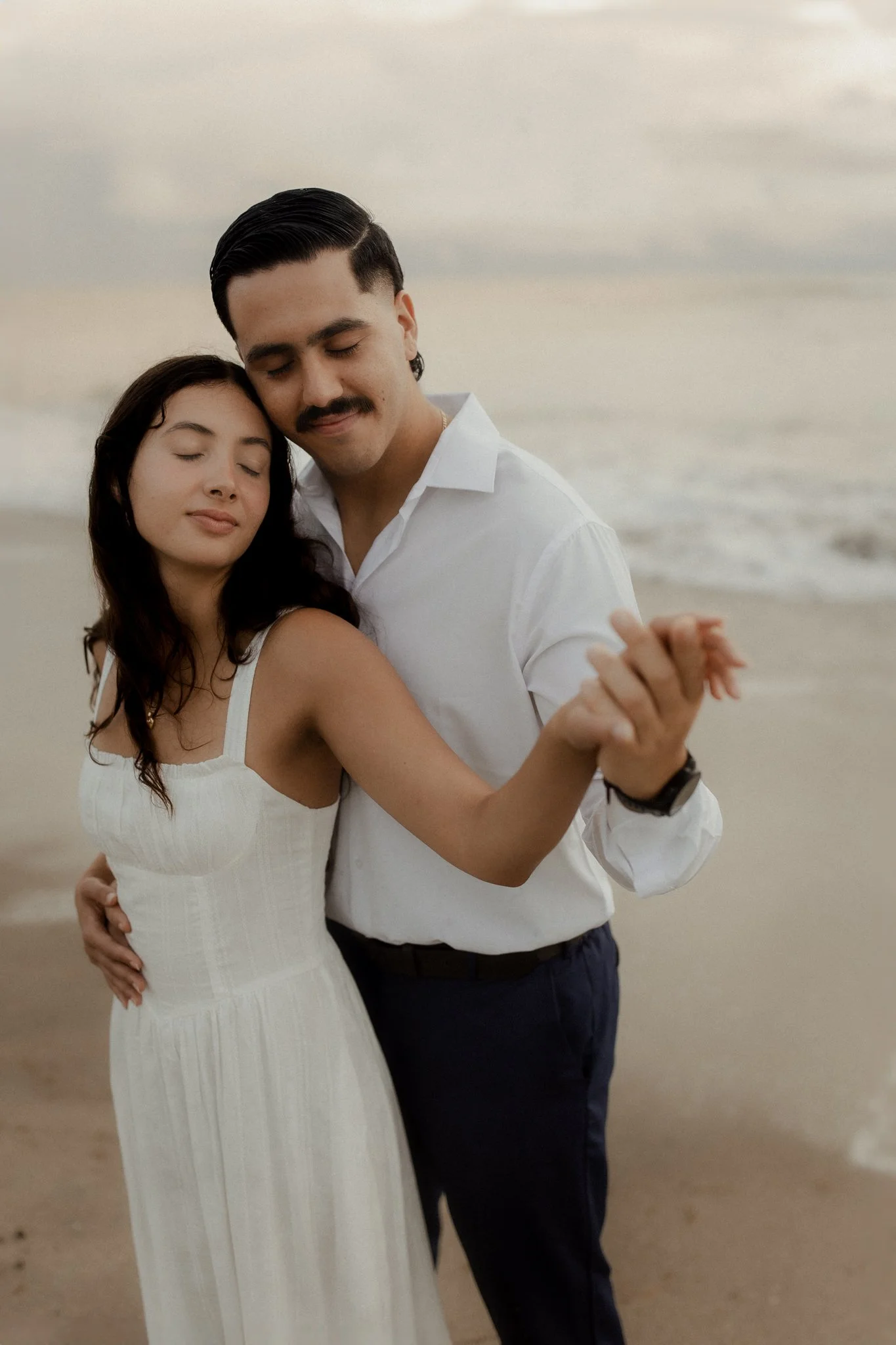 A couple dancing on the beach with eyes closed, embracing, during sunset or late afternoon.