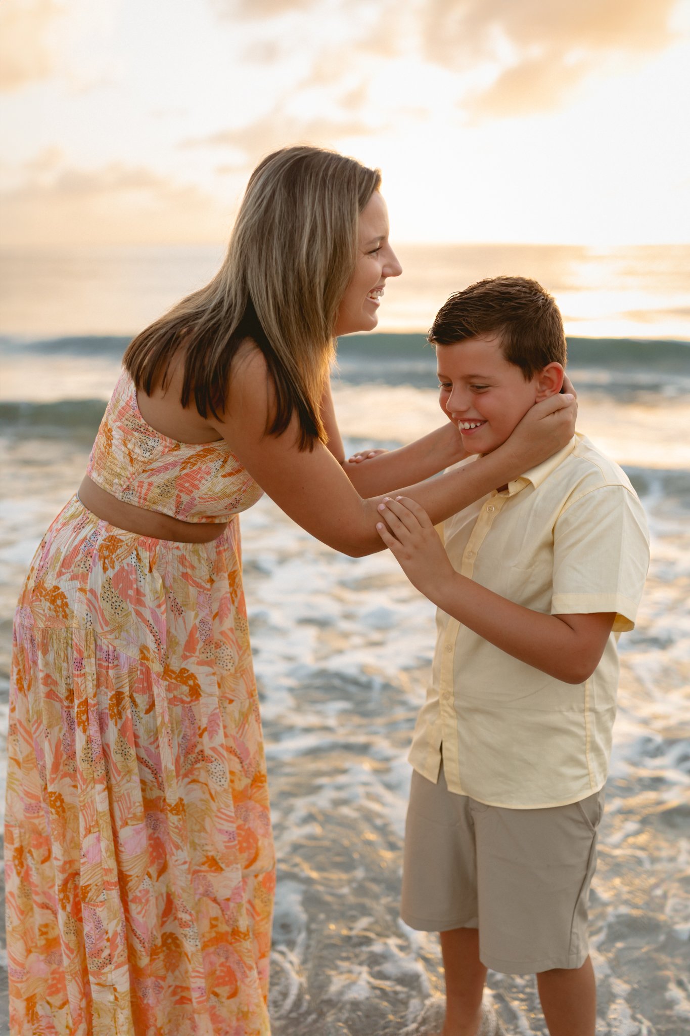 A woman and a boy smiling and playing at the beach during sunset, with the woman holding the boy's face gently.