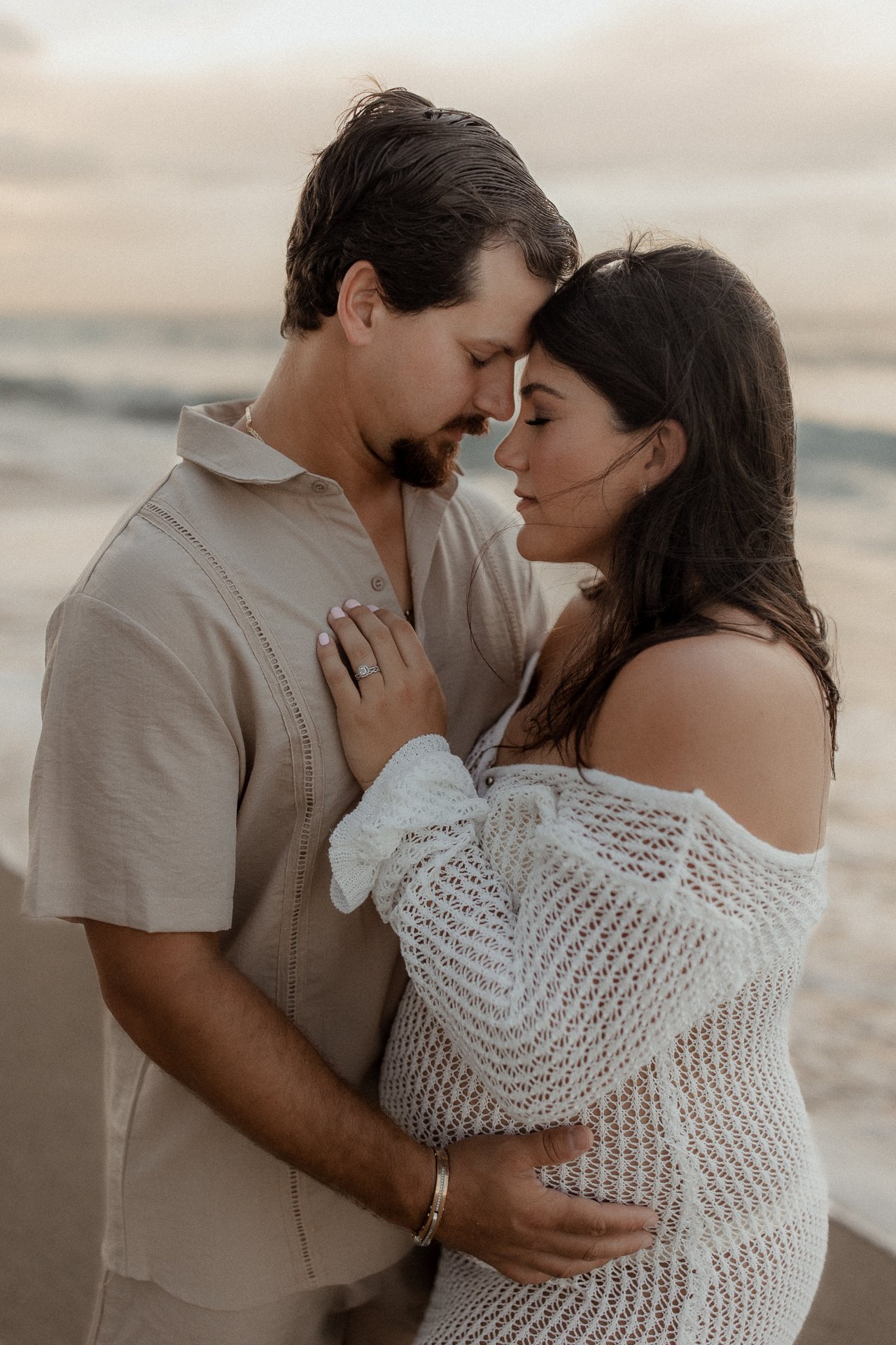 A couple embracing on the beach with their foreheads touching, eyes closed, during sunset. The woman is wearing a white crochet dress and an engagement ring, and the man is dressed in a beige shirt.