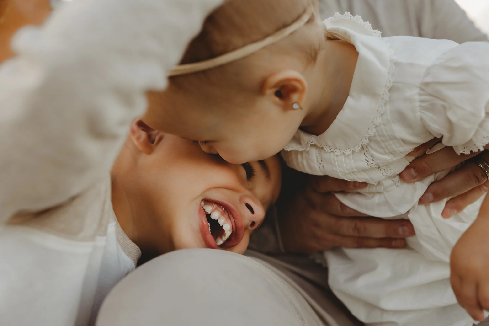 A woman lying down with a young girl and a baby girl. The woman and girl are smiling, and the girl is leaning over to kiss the baby.