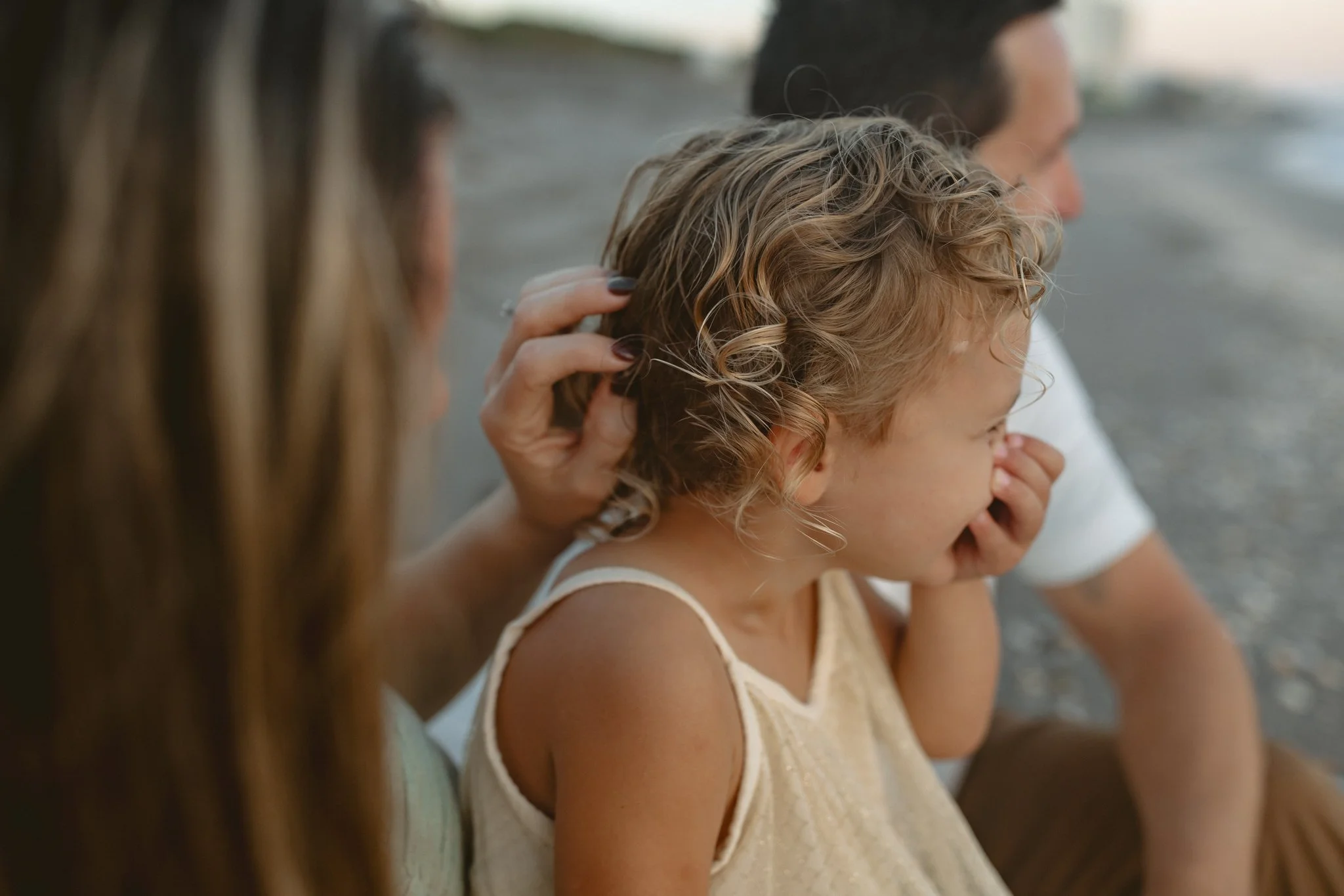 A family sitting on a beach, with a woman gently touching a young girl with curly hair, while a man watches in the background.
