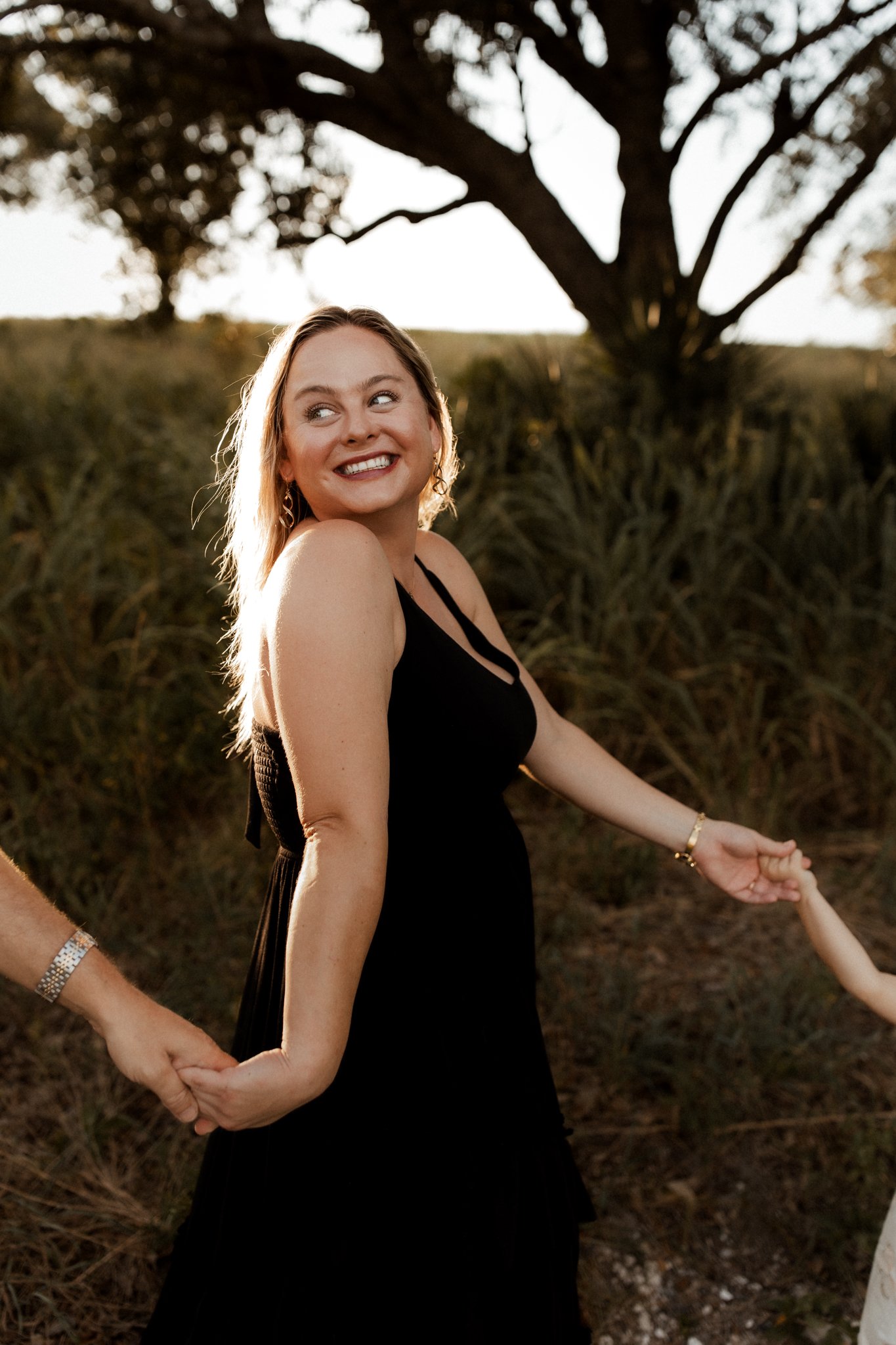 A woman in a black dress smiling and holding hands with others outdoors during sunset.