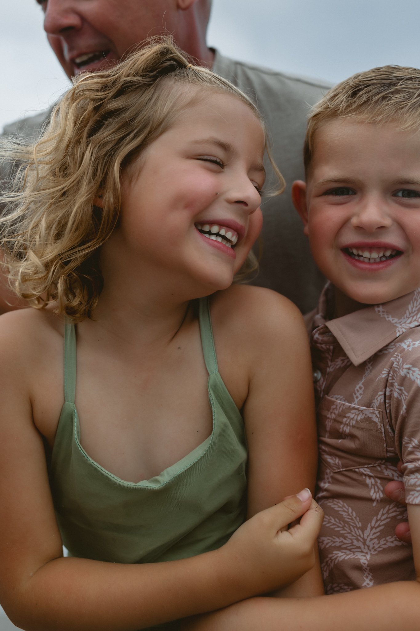 A close-up of a smiling girl with curly blonde hair next to a smiling boy with short blonde hair, with an adult partially visible behind them.